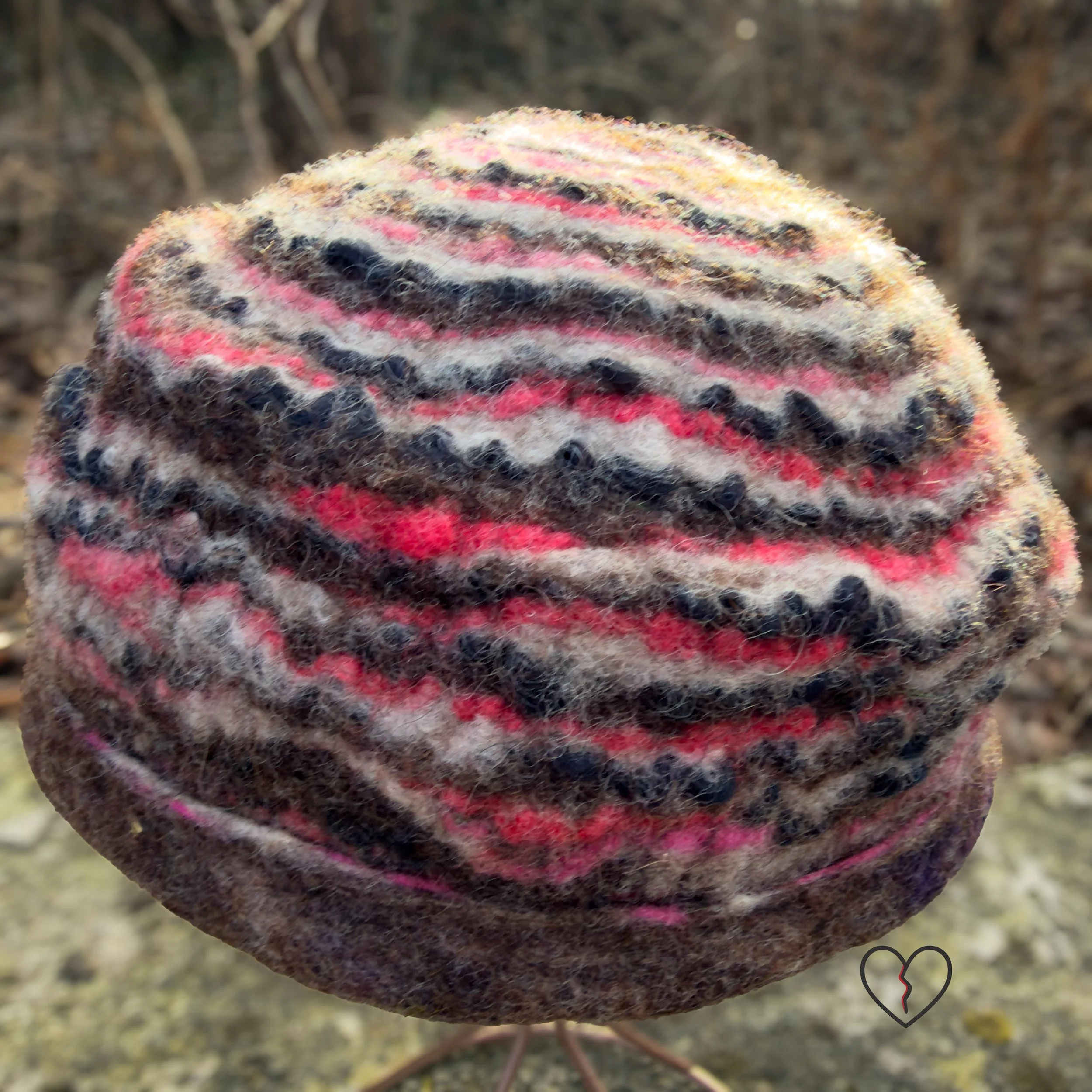 Side view of a sculptural wet-felted wool hat with undulating stripes and a soft structural fold, made from natural brown, white, and pink merino wool.
