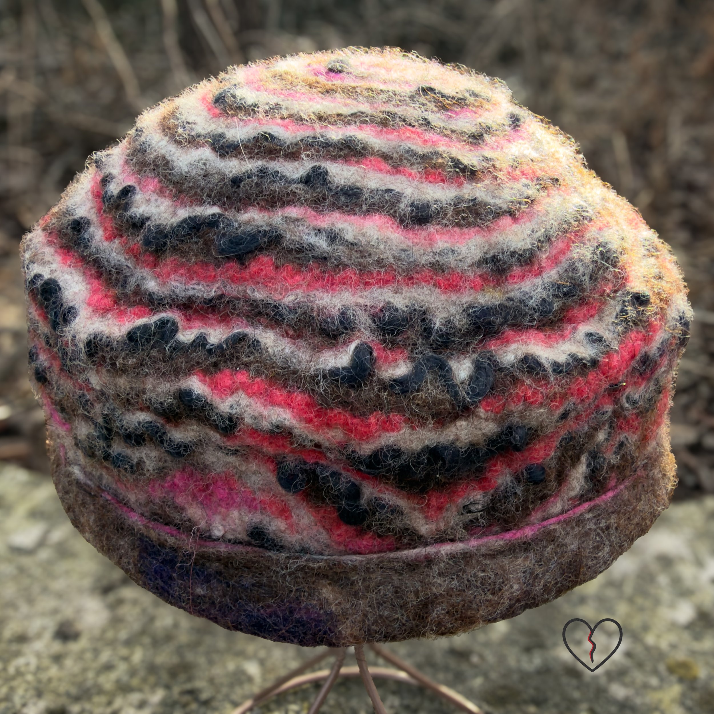 Front view of a one-of-a-kind hand-felted wool hat on a stand, showing rippling horizontal stripes and a folded brim in natural wool tones with pink accents.