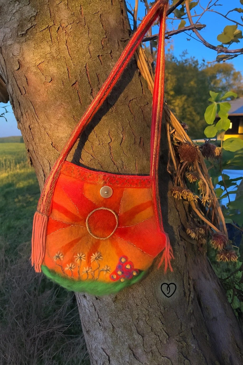 Handmade felted wool shoulder bag featuring a radiant sun motif with embroidered rays, needle-felted flowers, and a crocheted strap, photographed outdoors hanging from a tree.