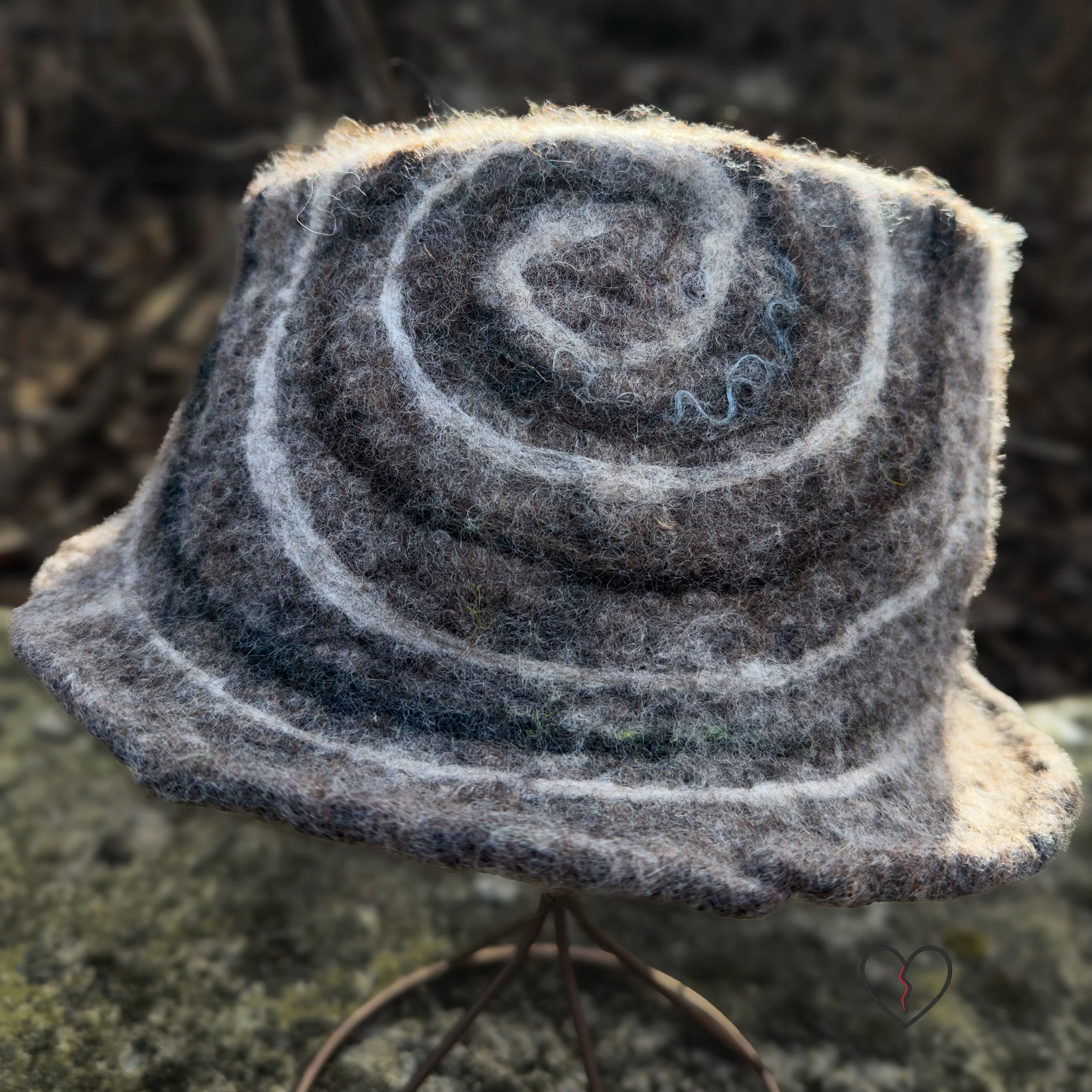 Hand-felted wool fedora hat in natural brown tones with a white and dark brown spiral design, sculptural crown, and short brim, photographed outdoors.