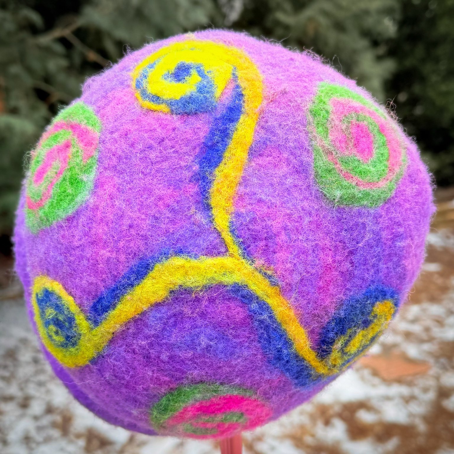 Top view of handmade purple felted wool hat featuring a triskele symbol with blue and yellow arms and surrounding spiral motifs