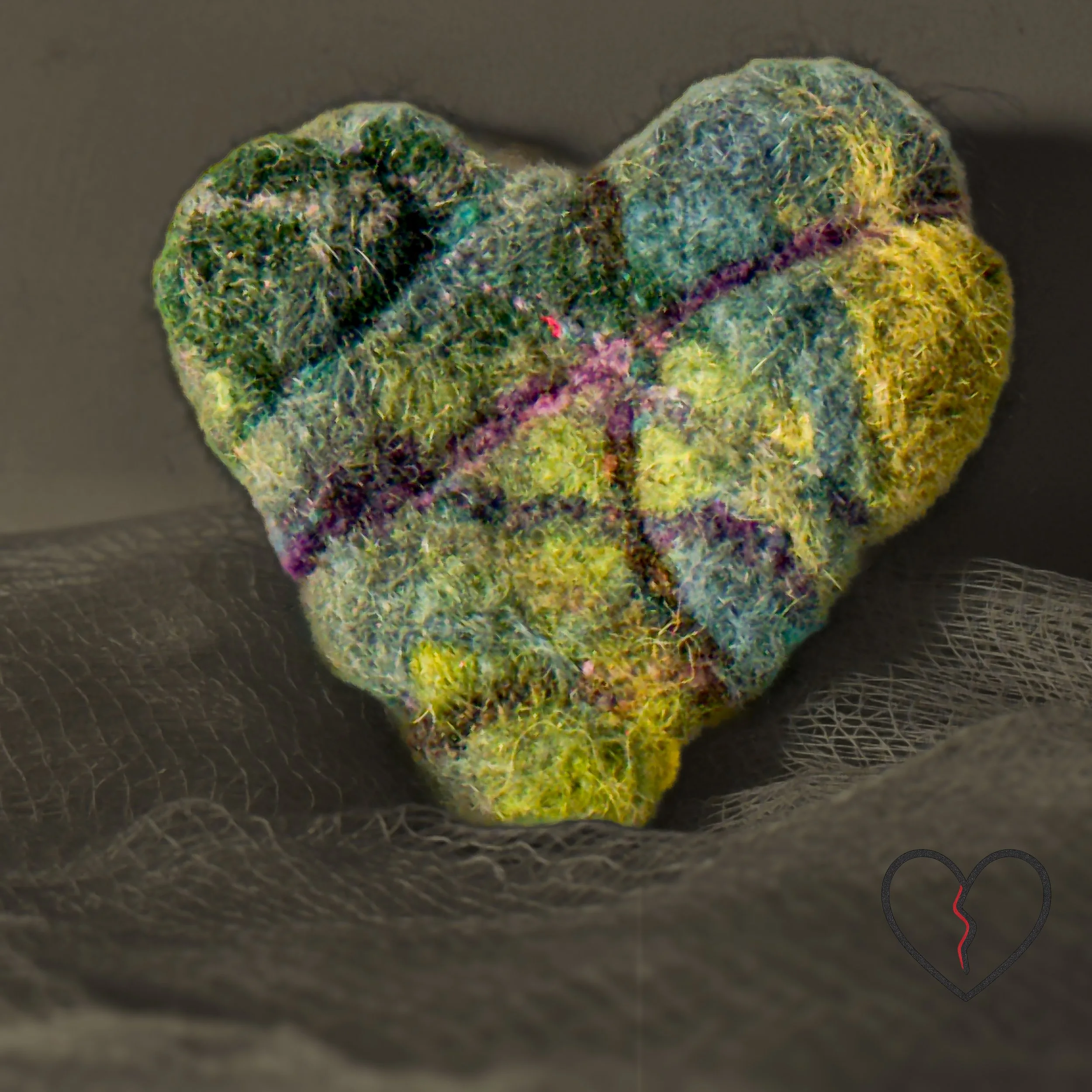 Small wool heart art pin photographed on soft gray fabric, highlighting texture and organic color variations