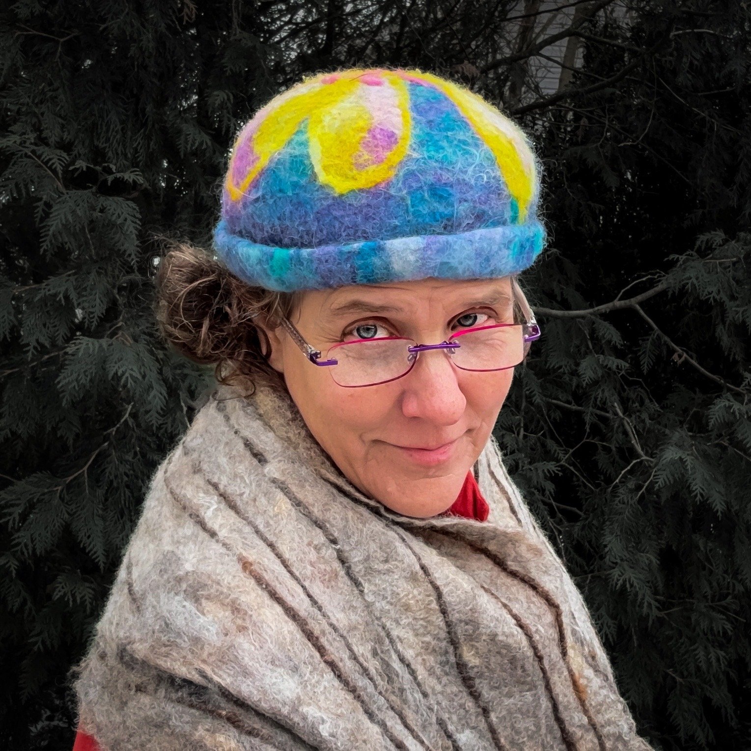 Artist wearing a hand-felted wool hat with a blue angora-blended base and a colorful merino flower design, photographed outdoors against dark evergreen trees in winter.