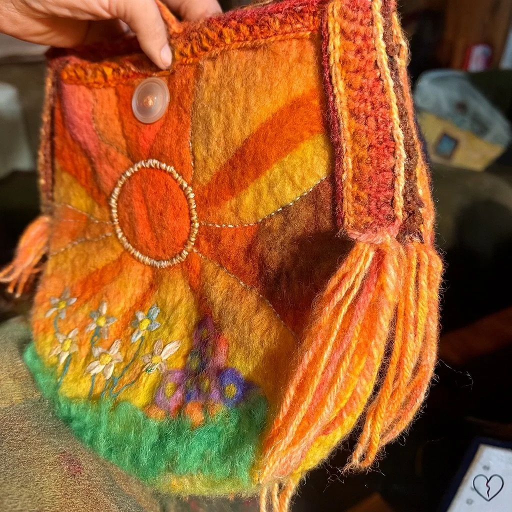 Hand felted merino wool purse with a long crocheted shoulder strap, embroidered sun design, and needle-felted floral garden imagery, shown hanging from a tree trunk.