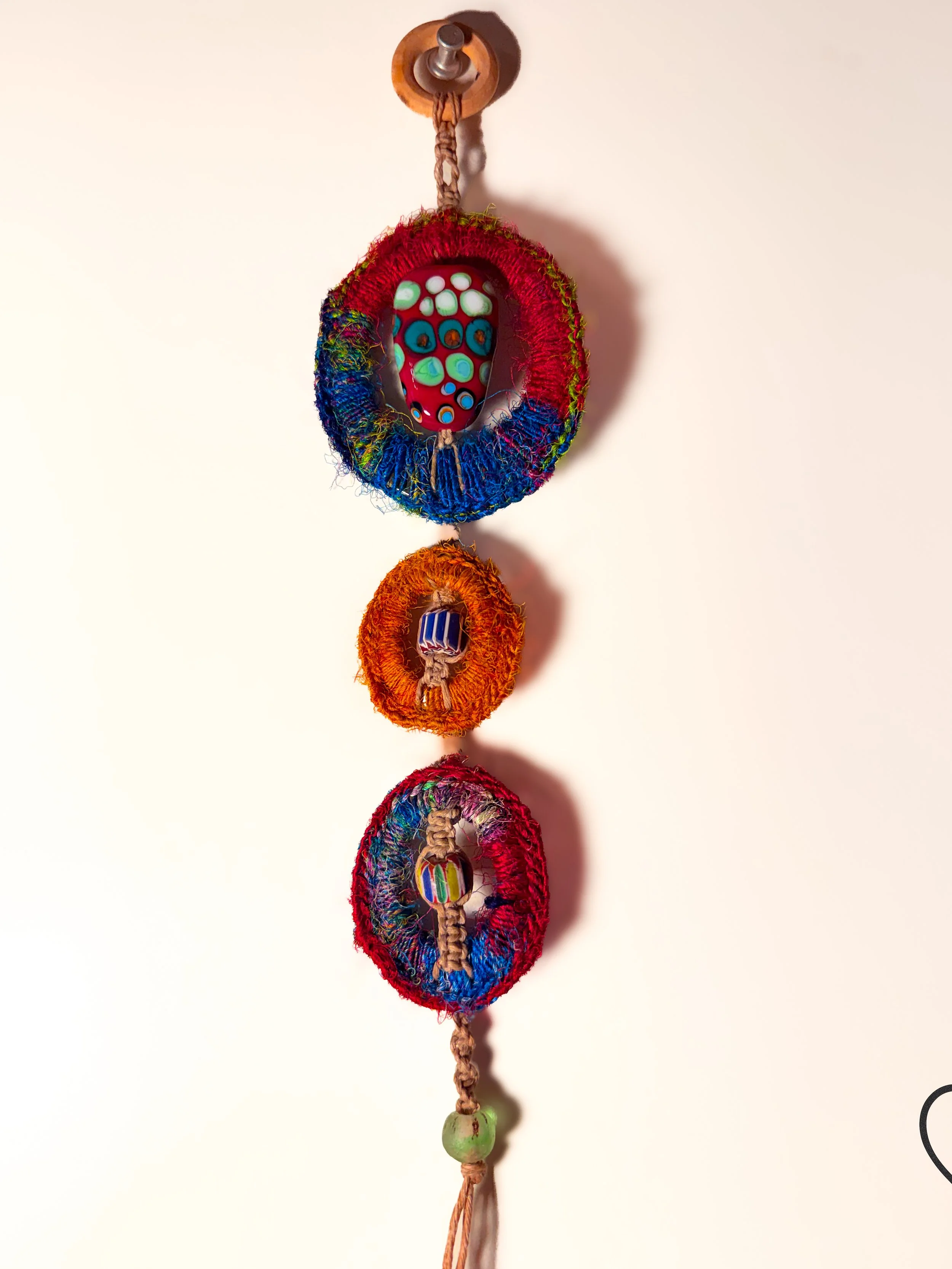 Trinity Weaver, 2024, by Kristi Yapp is a three circle Dreamcatcher made from reclaimed metal, sari silk, hemp, and glass beads.