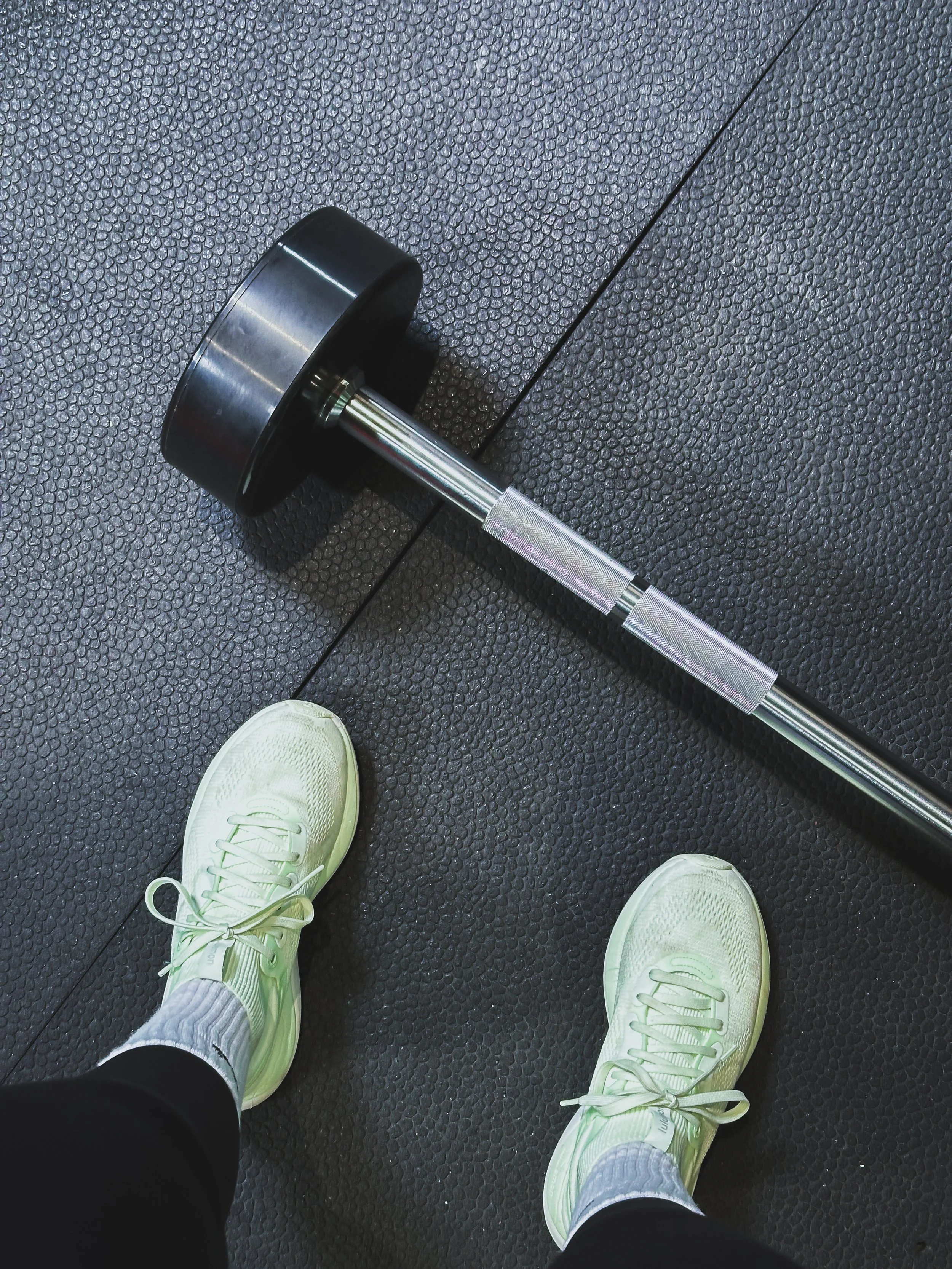 A person standing on a black gym floor with white athletic shoes, wearing black pants and grey socks, next to a black self-weights barbell with a large black weight plate on a textured rubber mat.