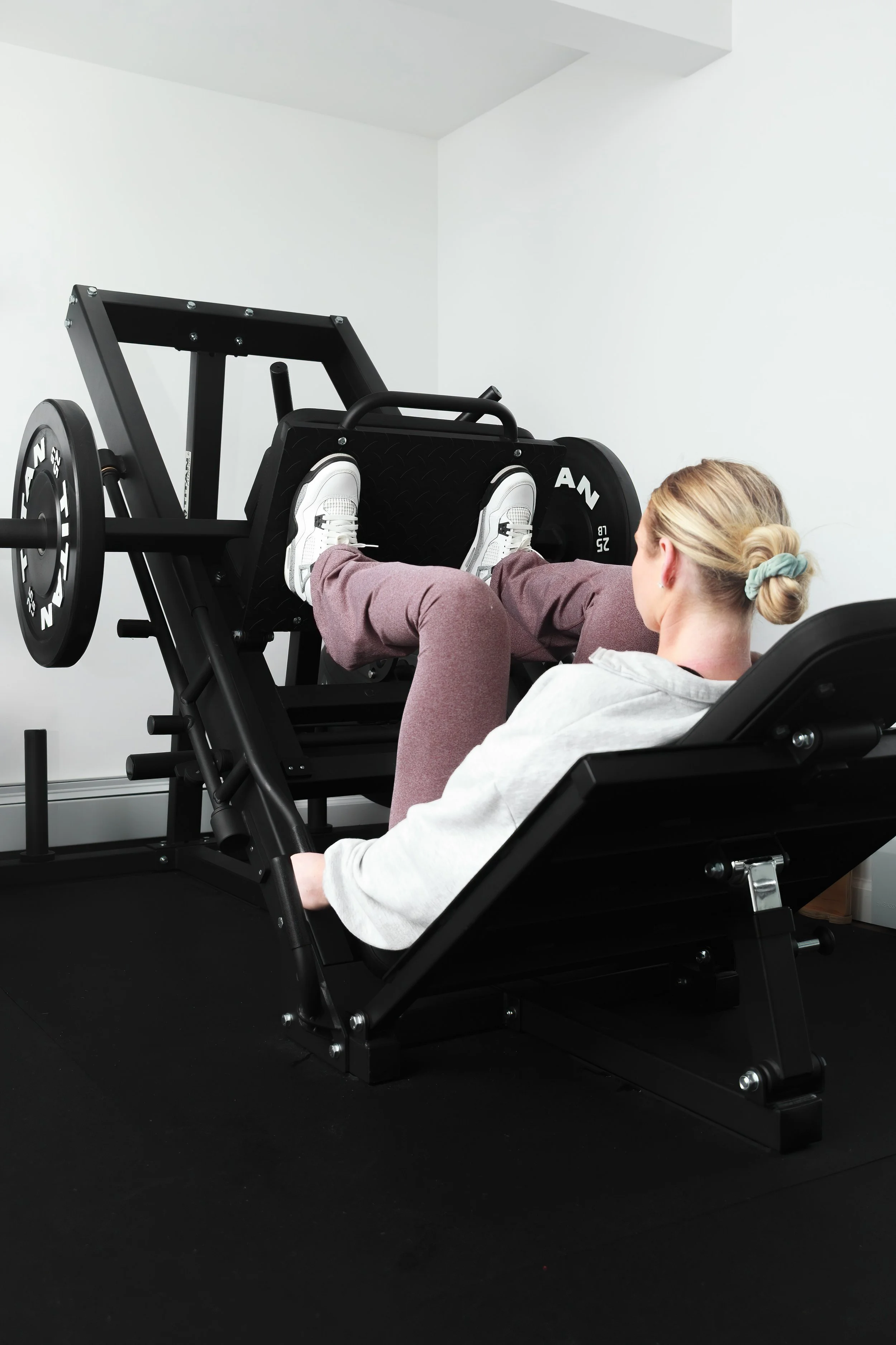 A woman in comfortable workout clothes doing a leg exercise on a specialized black fitness machine in a bright, clean room.