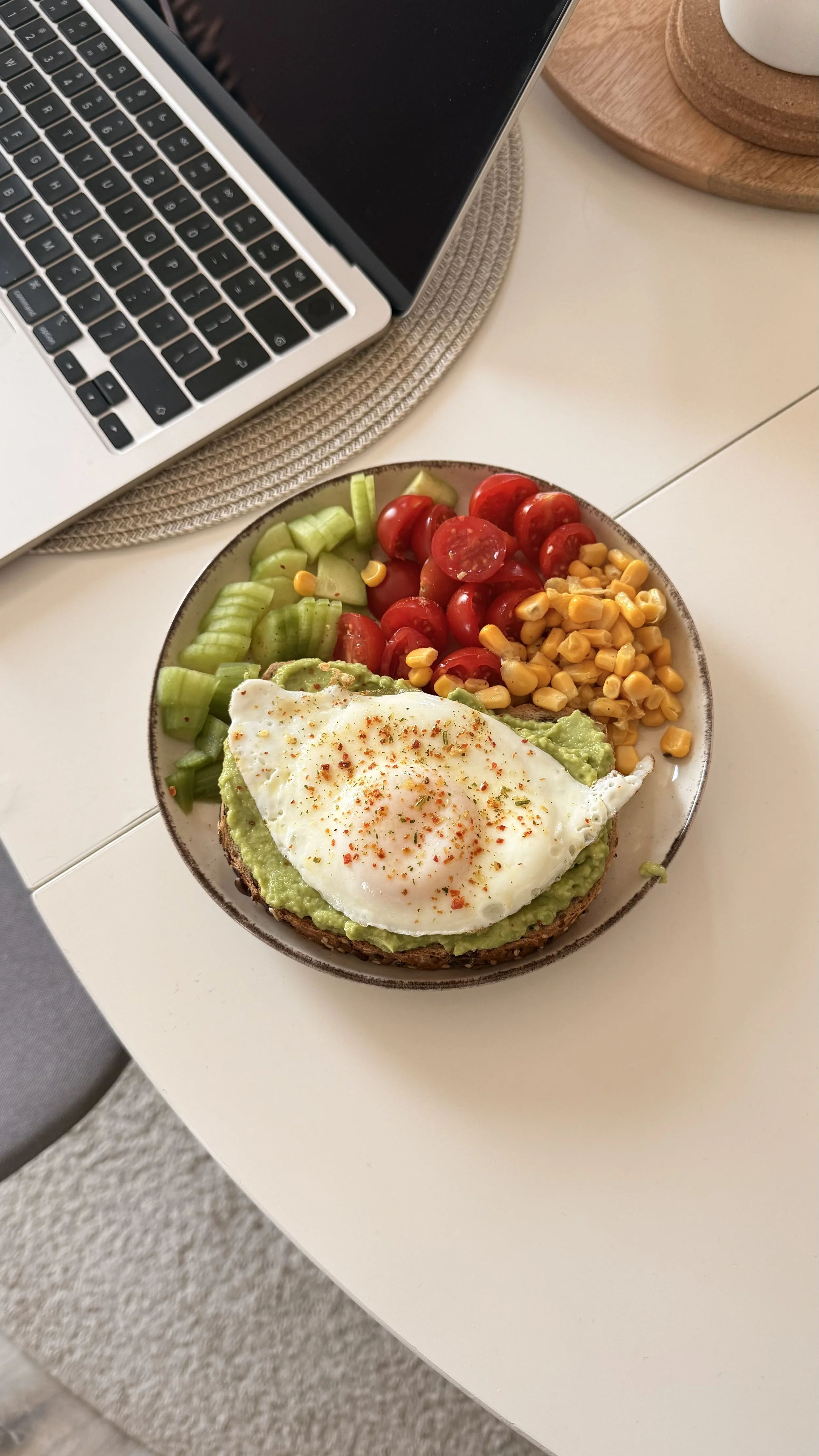 A bowl of avocado toast topped with a fried egg, garnished with red pepper flakes, alongside cherry tomatoes, celery slices, and corn, on a white table with a laptop nearby.