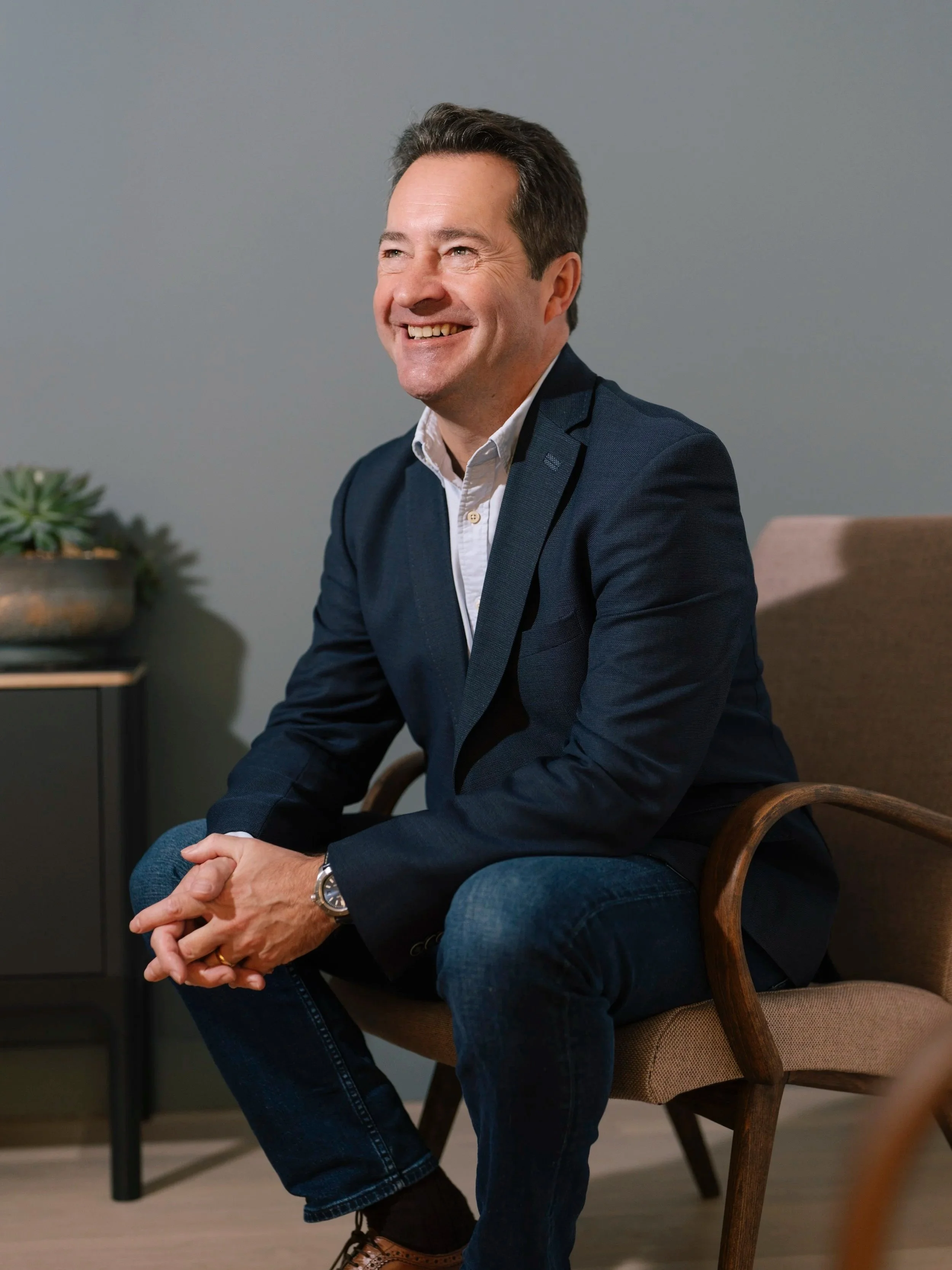 Steve Currie, Chairman, sitting on a chair, smiling in a modern, casual office setting.