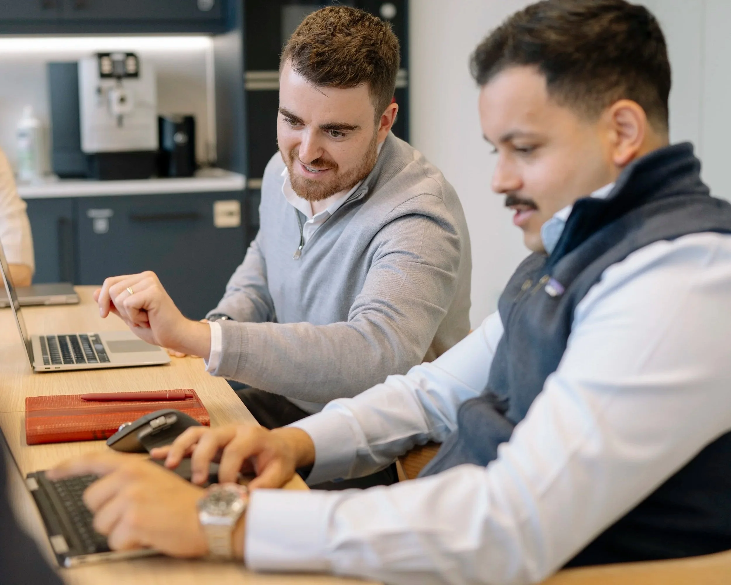 Two men working together at a conference table with laptops in an office setting.