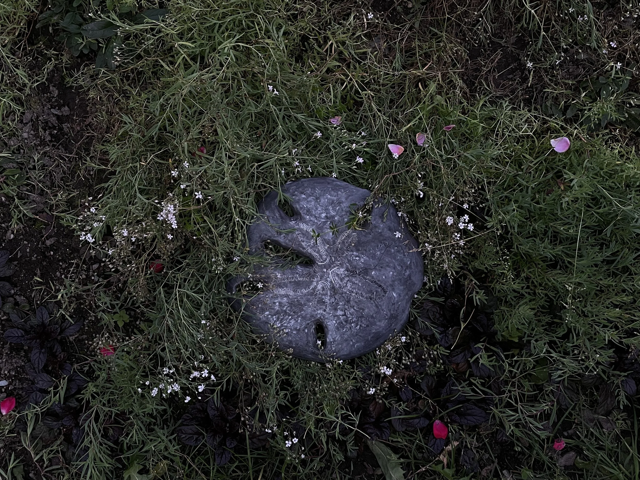A round metallic sculpture resembling a face, surrounded by green plants, small white flowers, and pink petals.
