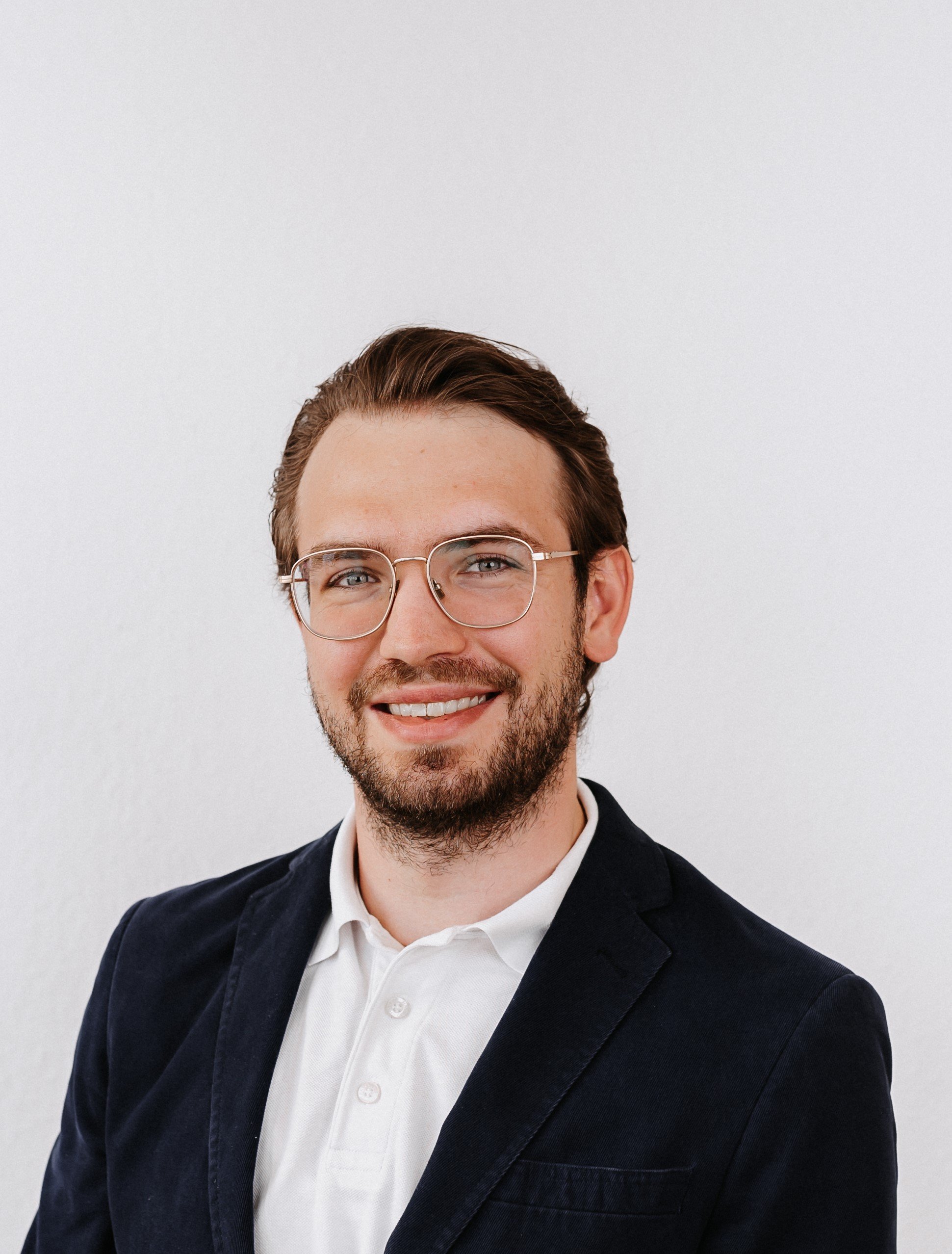 Porträt eines lachenden jungen Mannes mit Brille, dunklem Jackett und weißem Hemd vor einem weißen Hintergrund.