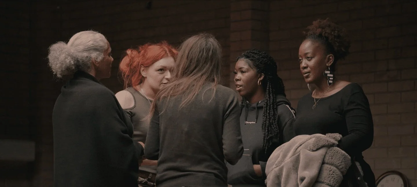 Five women engaged in conversation indoors with a brick wall in the background.