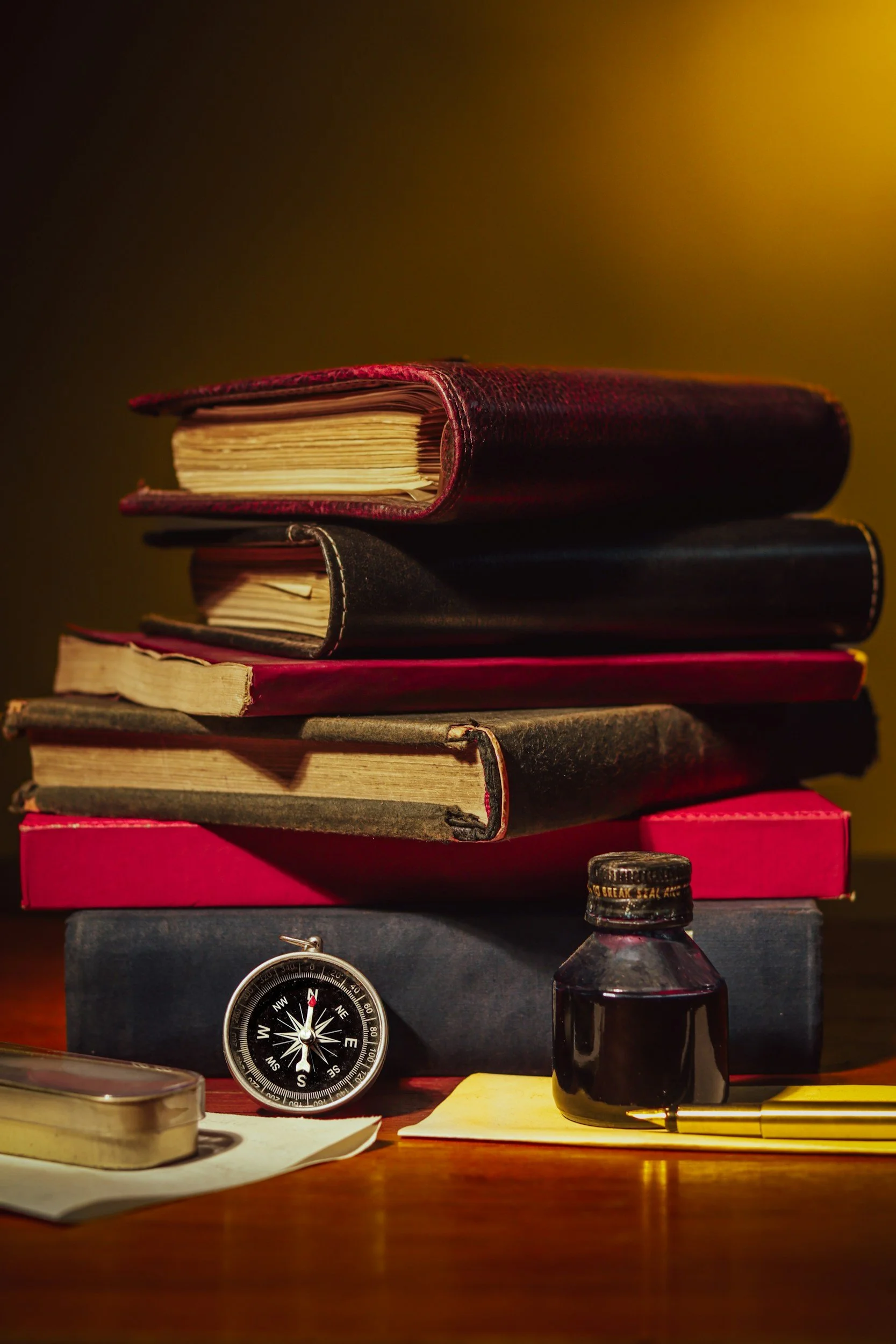 Stacked books on a desk, with a compass, a small bottle, a pen, and papers in the foreground.