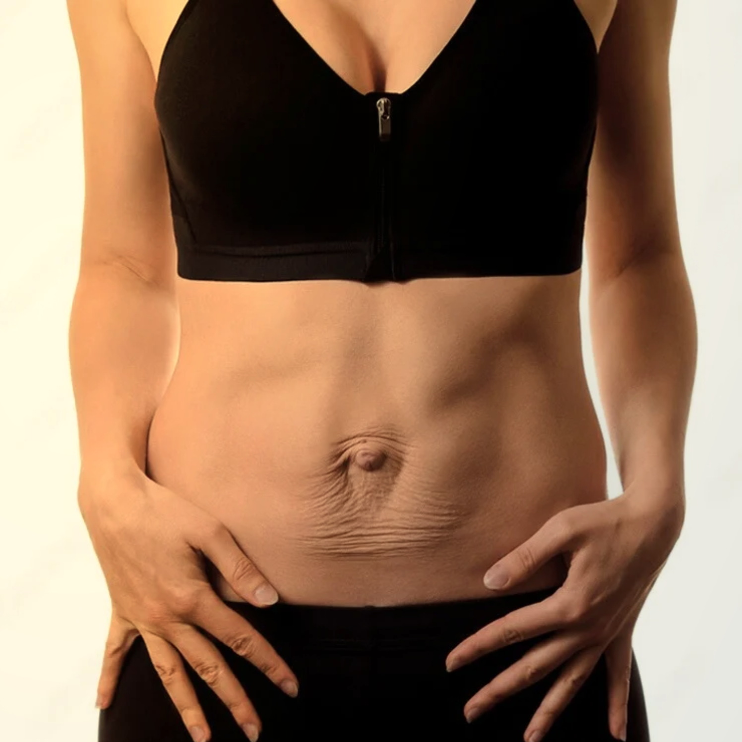 Mulher com roupas pretas, vestindo top com zíper na frente, mãos nas laterais da barriga, destacando o abdômen e o umbigo.