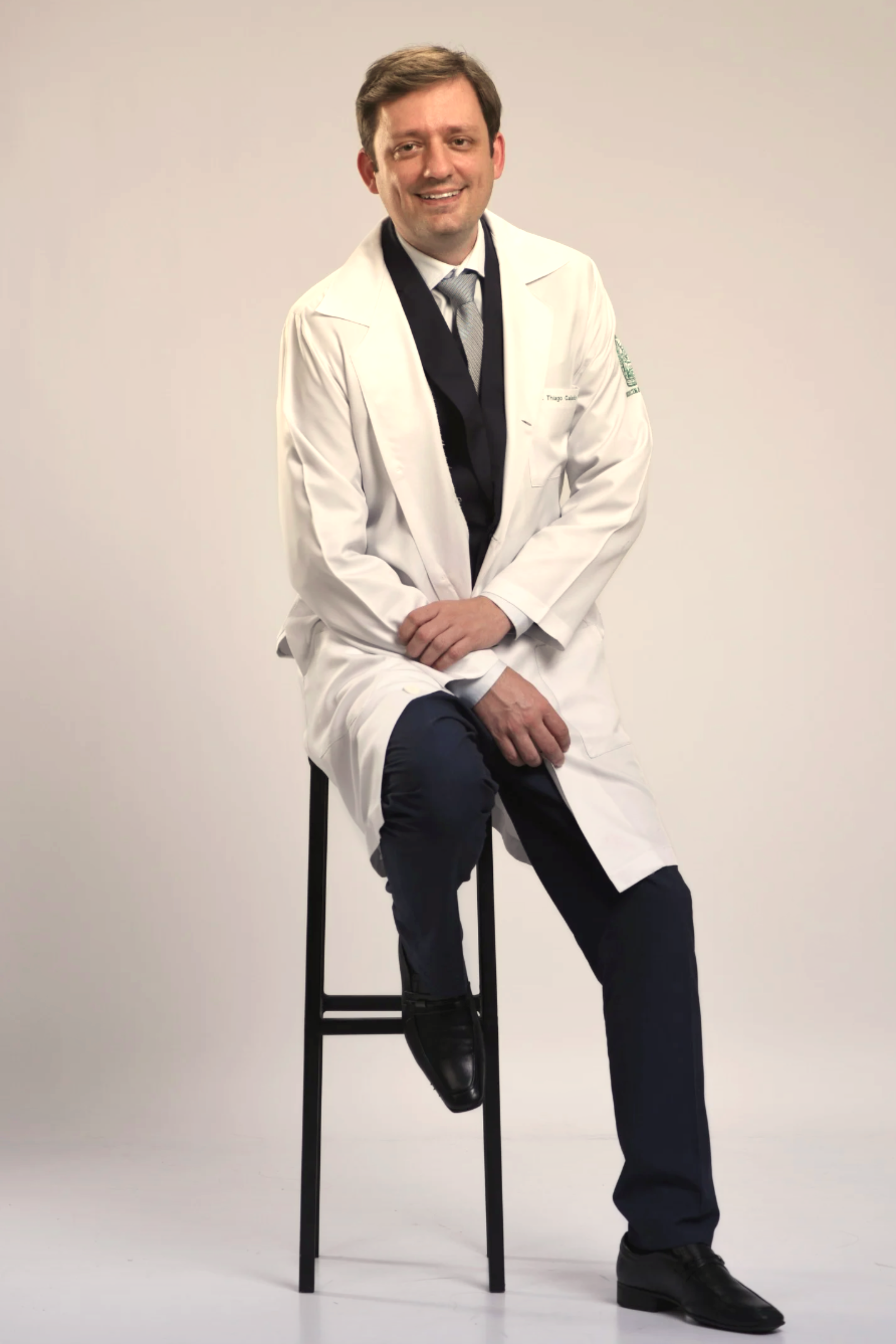 Homem sorridente, de terno e jaleco branco, sentado em um banquinho contra fundo branco.