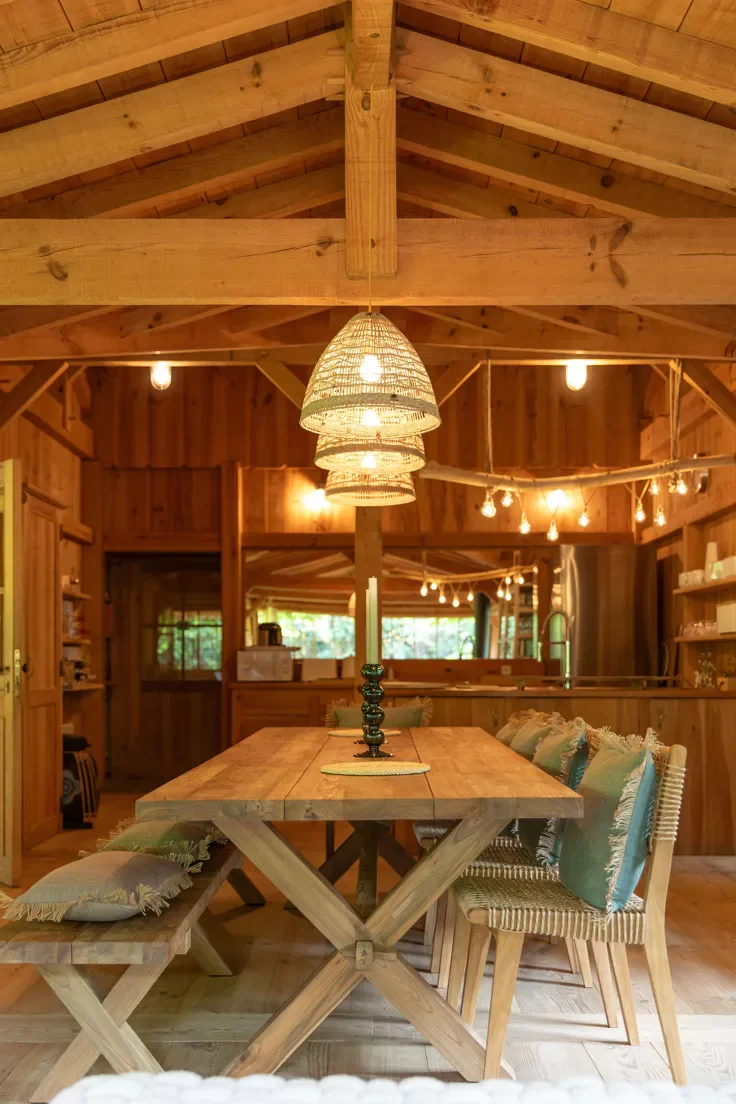 Intérieur en bois d'une salle à manger avec une table en bois, chaises avec coussins, et un centre de table, éclairage en suspended avec des lampes en tissu.