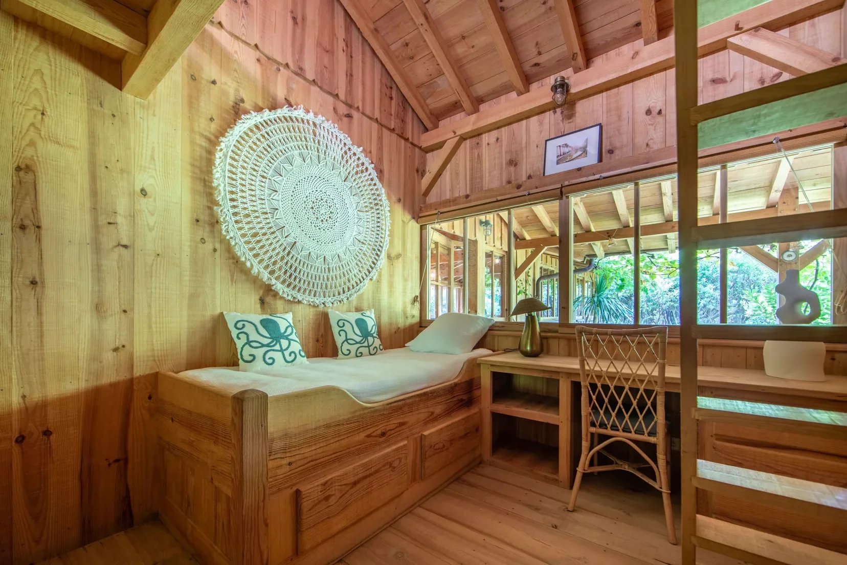 Intérieur d'une cabane en bois avec un lit, des coussins en forme de pieuvre, une lampe en or, une table en bois, une chaise en osier et une grande fenêtre regardant sur un jardin