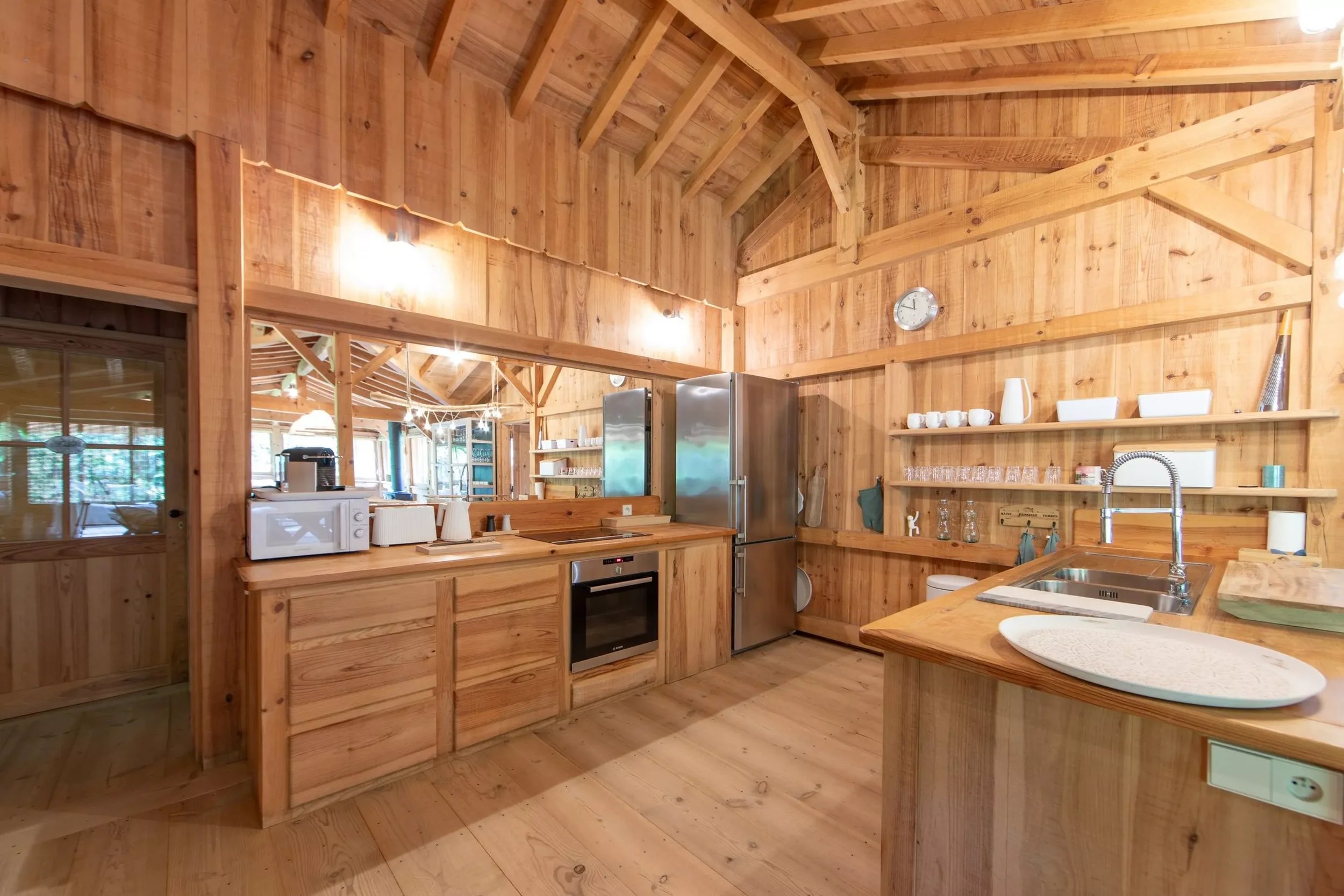 Cuisine en bois avec un réfrigérateur, micro-ondes, évier, étagères avec vaisselle, tableau mural et ouverture sur une salle de séjour lumineuse.
