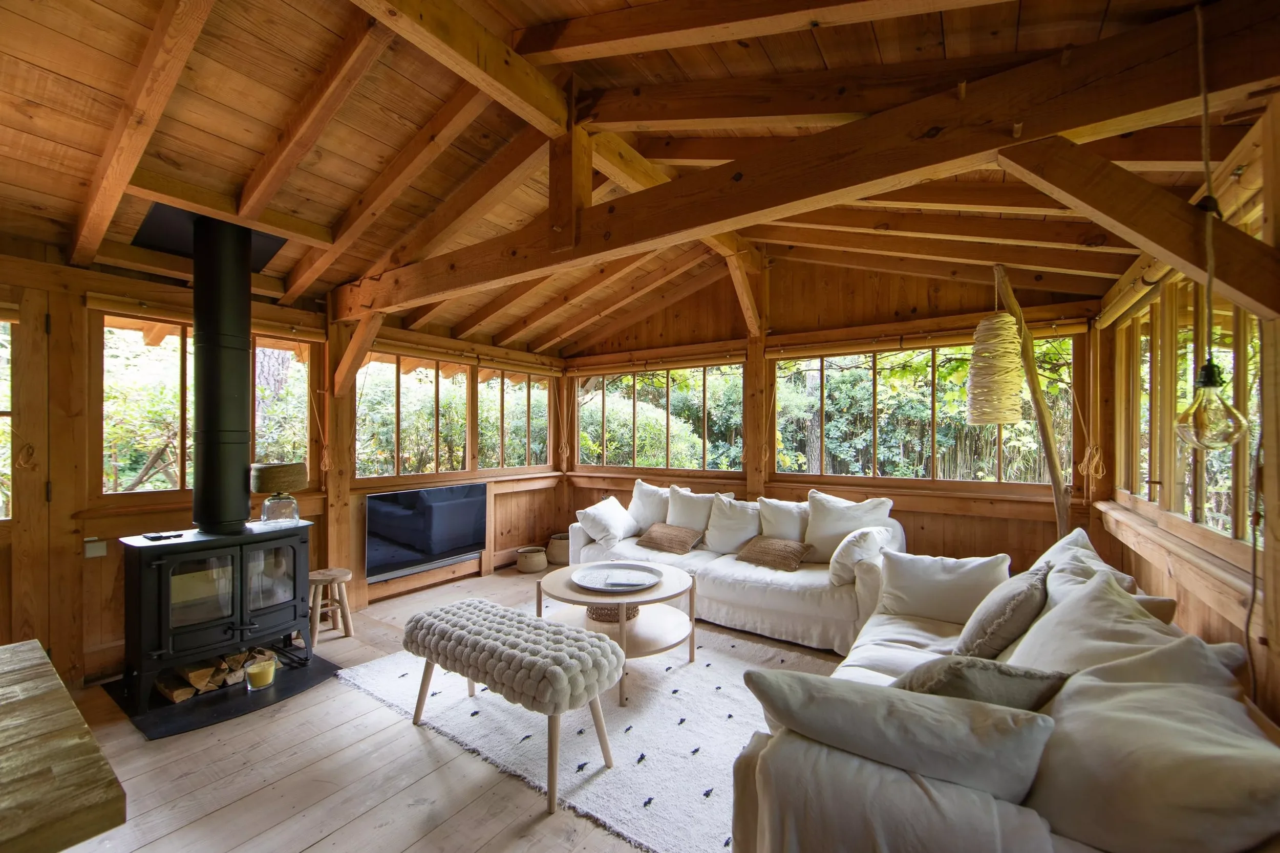 Salon en bois avec canapés blancs, cheminée à insert, fenêtres donnant sur la forêt, tapis blanc, table basse ronde, pouf en laine, lumière suspendue, ambiance chaleureuse et naturelle.