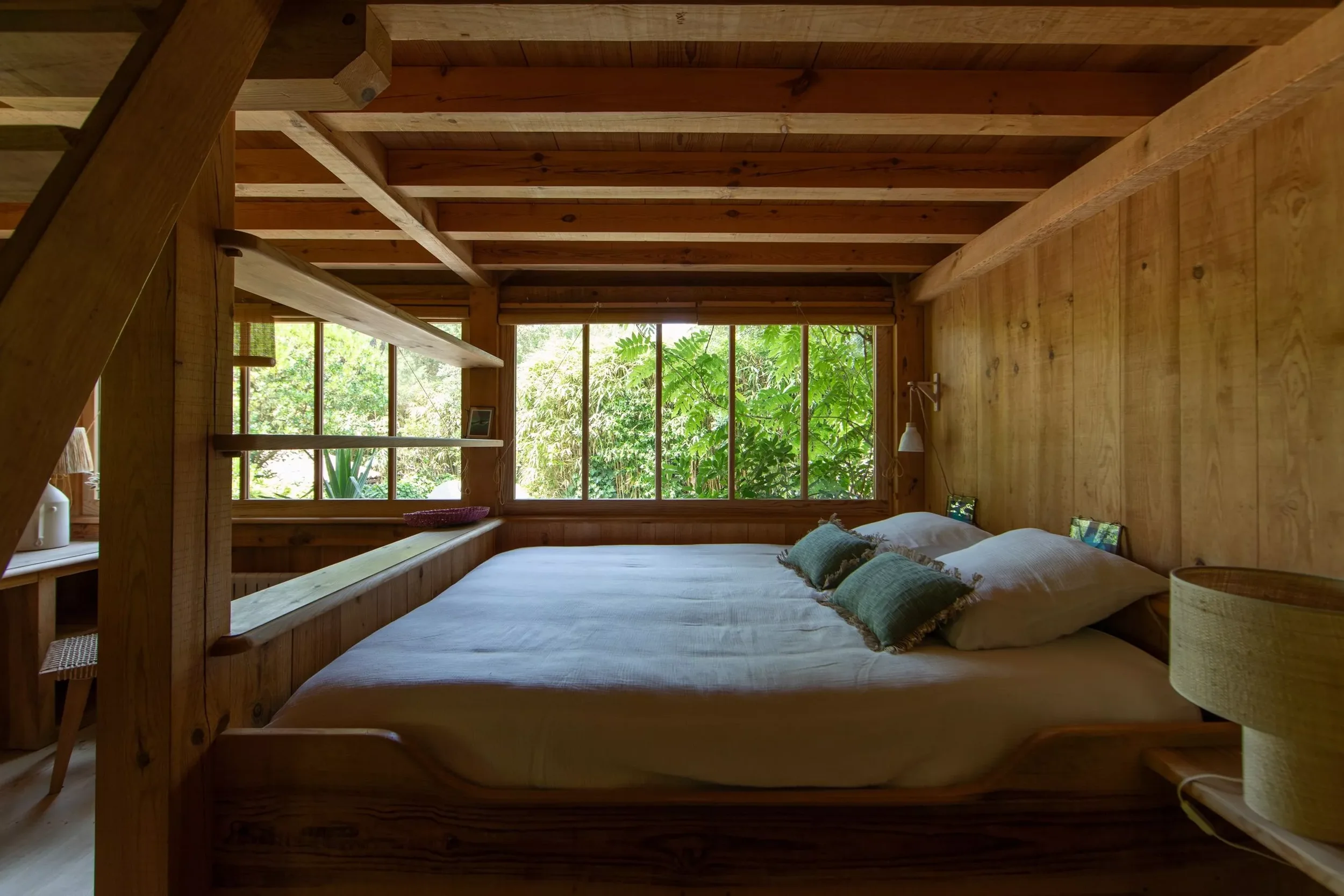 Intérieur d'une chambre en bois avec un grand lit, coussins verts et fenêtre donnant sur un jardin verdoyant.