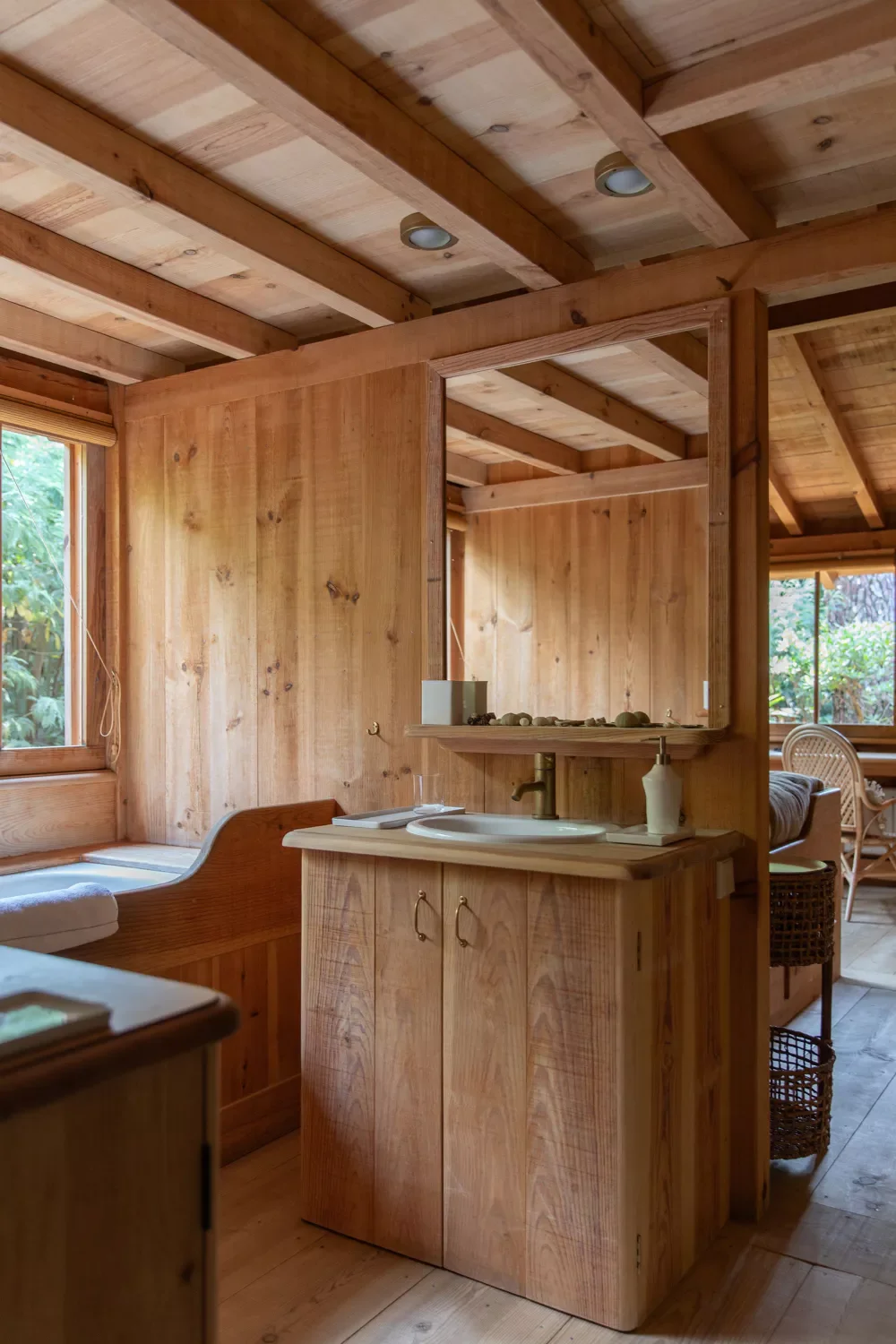 Intérieur d'une pièce en bois avec un petit lavabo, un miroir, une fenêtre laissant entrer la lumière naturelle, et un coin salon avec un banc en bois et un coussin.