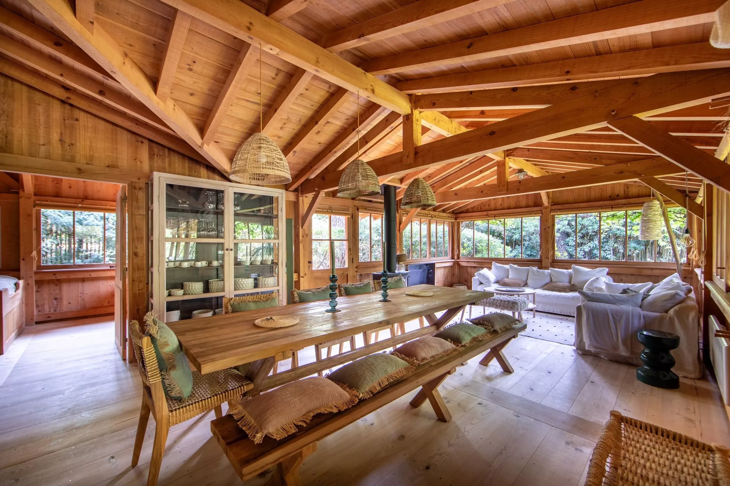 Intérieur d'une maison en bois avec un plafond en bois, un grand lit en coin et une grande table en bois avec des chaises et des coussins, grandes fenêtres laissant entrer de la lumière naturelle