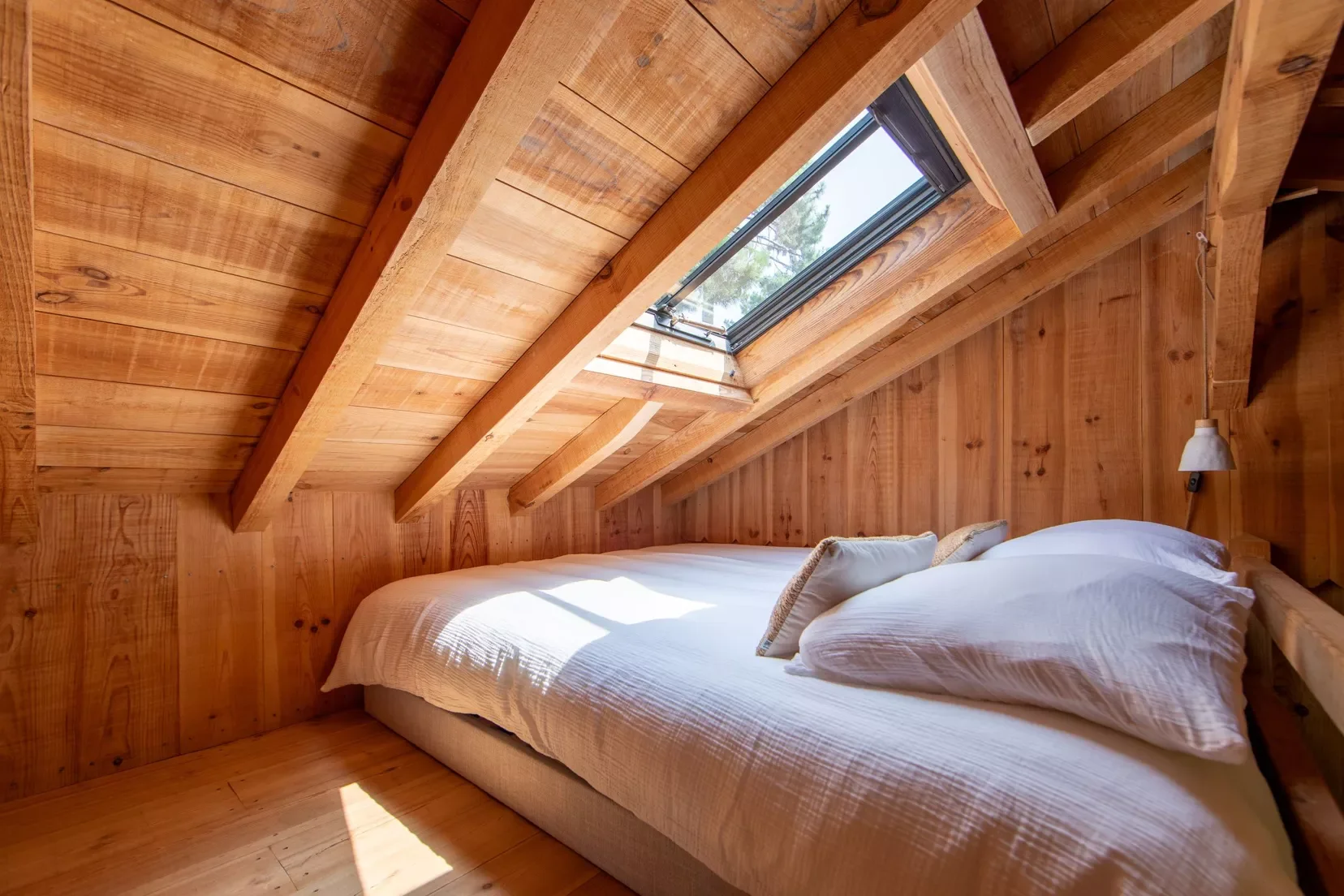 Chambre sous un toit en bois avec un lit, un mur en bois, un plafond en pente, et une fenêtre de toit laissant entrer la lumière naturelle.