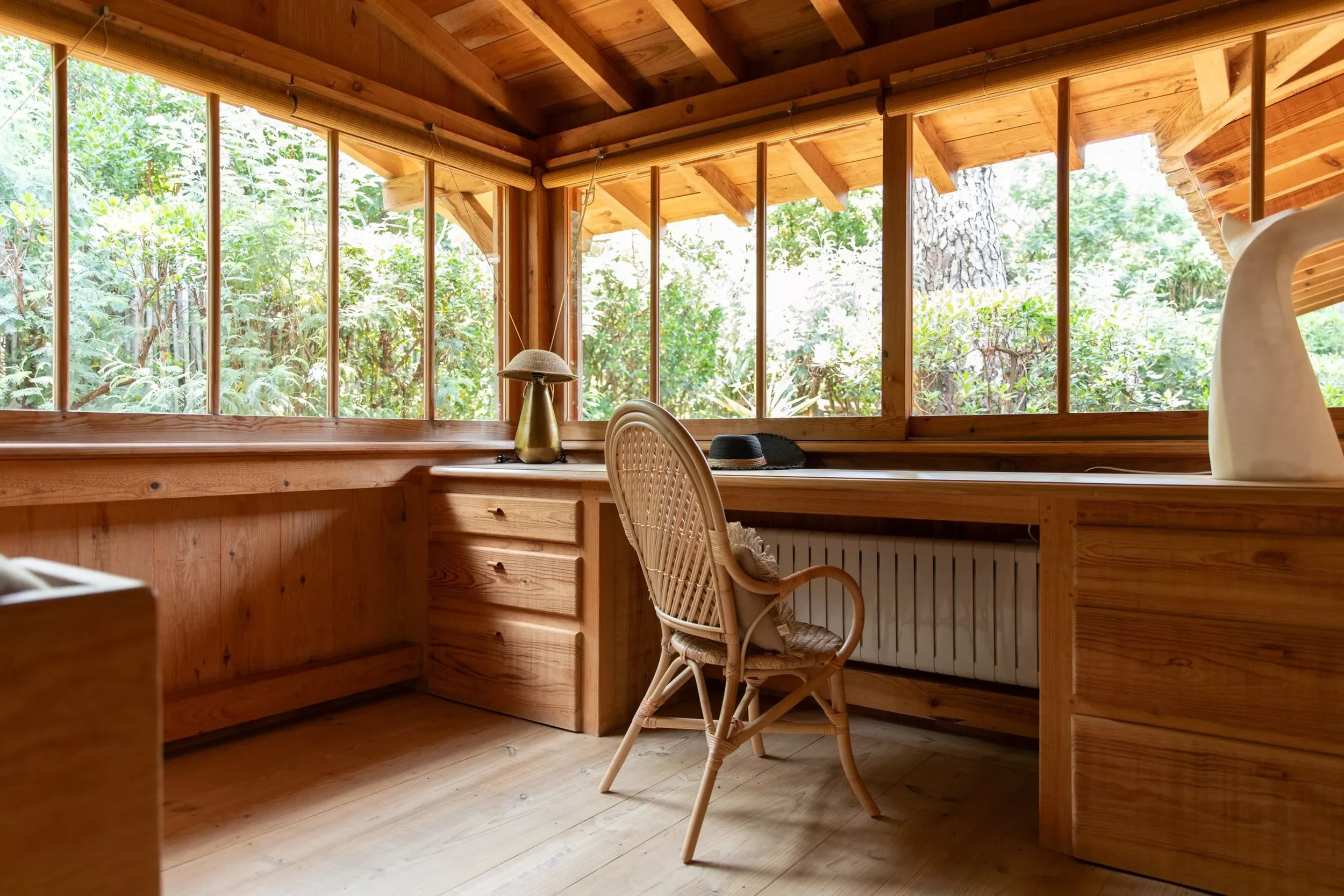 Espace de travail en bois avec bureau, chaise en rotin, lampe, chapeau noir, et décoration, vue sur la nature à travers de grandes fenêtres.