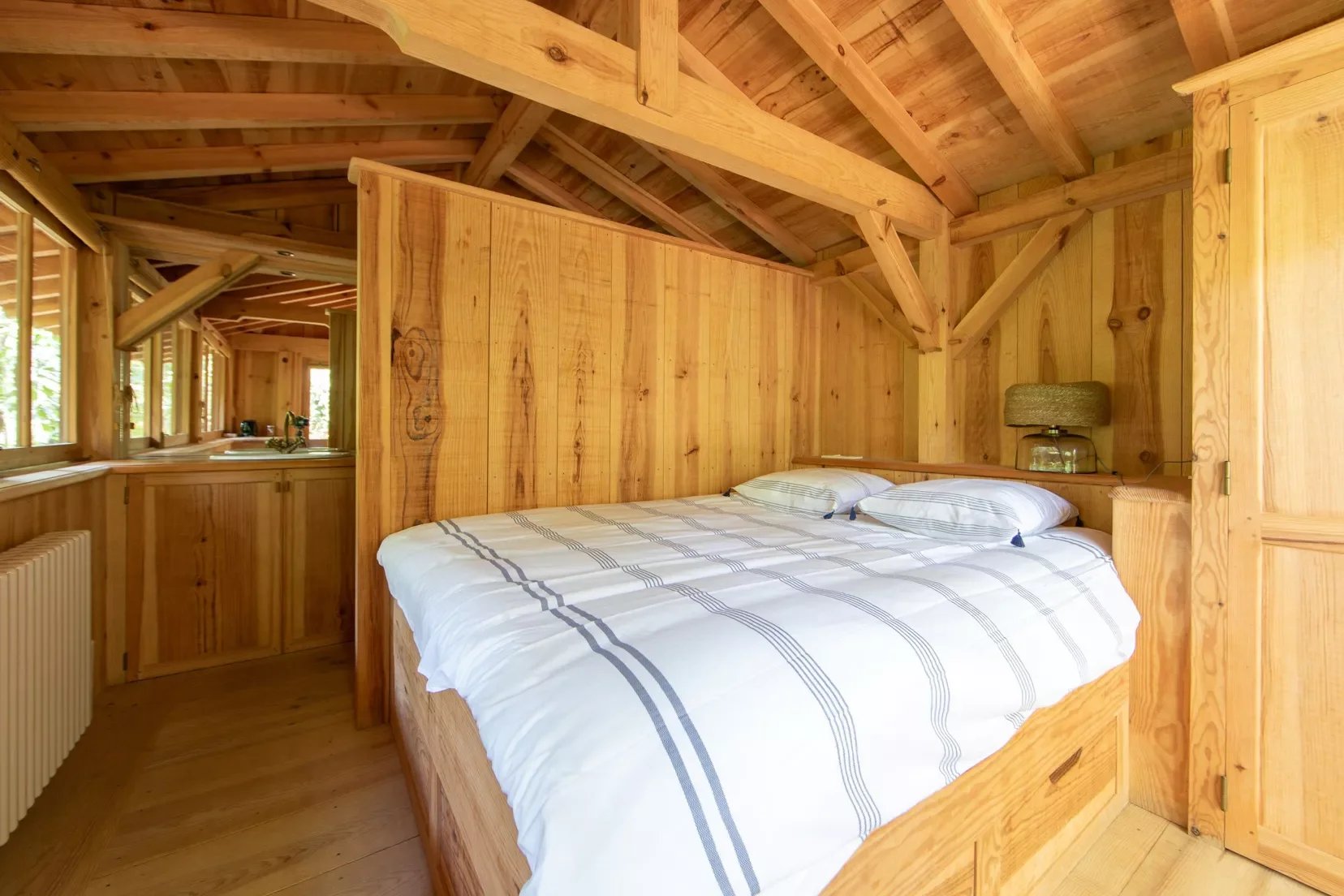Chambre en bois avec un lit double, fenêtres, et une lampe. La structure en bois est visible au plafond et aux murs.