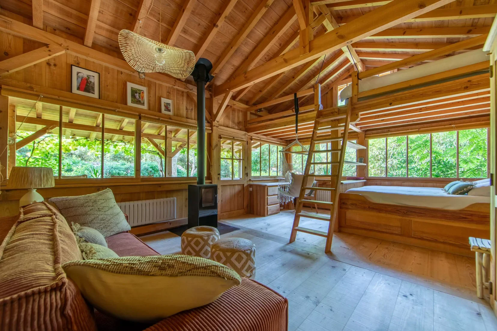 Intérieur d'une cabane en bois avec canapé, lit, grande fenêtre, échelle, fauteuil suspendu et cheminée