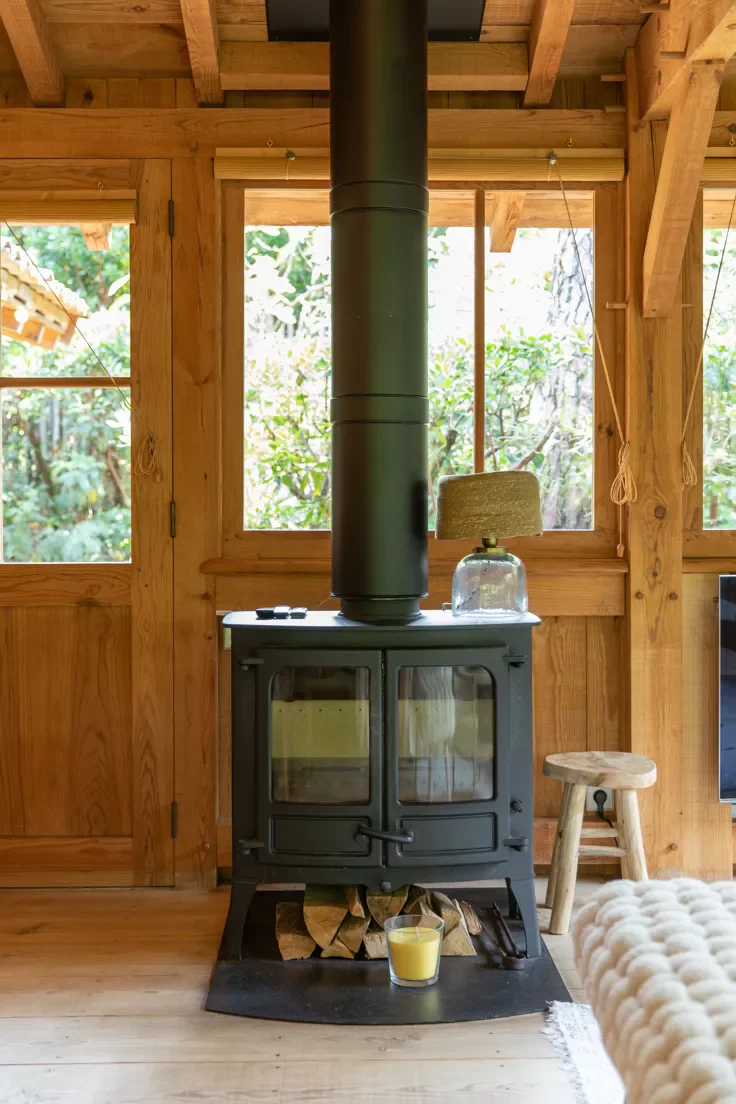 Intérieur en bois avec une cheminée en métal noire, associé à une cheminée en fonte, une lumière de bougie, un petit tabouret en bois, et un décor rustique.
