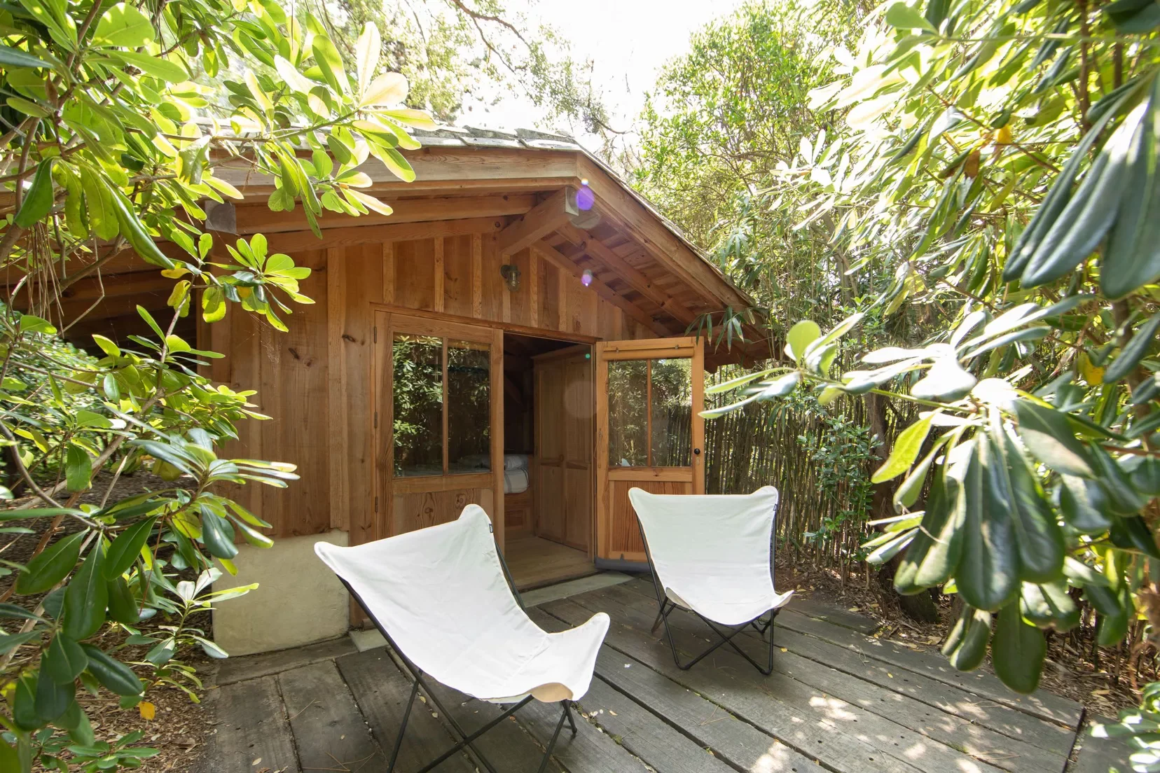 Petite cabine en bois entourée de végétation, avec deux chaises pliantes blanches sur une plateforme en bois.