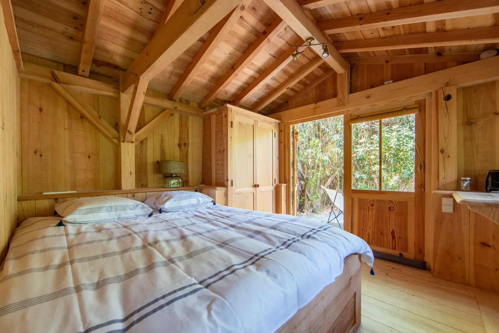 Chambre en bois avec lit double, fenêtre ouverte donnant sur la nature, meubles en bois, lampe, petite table et radio.
