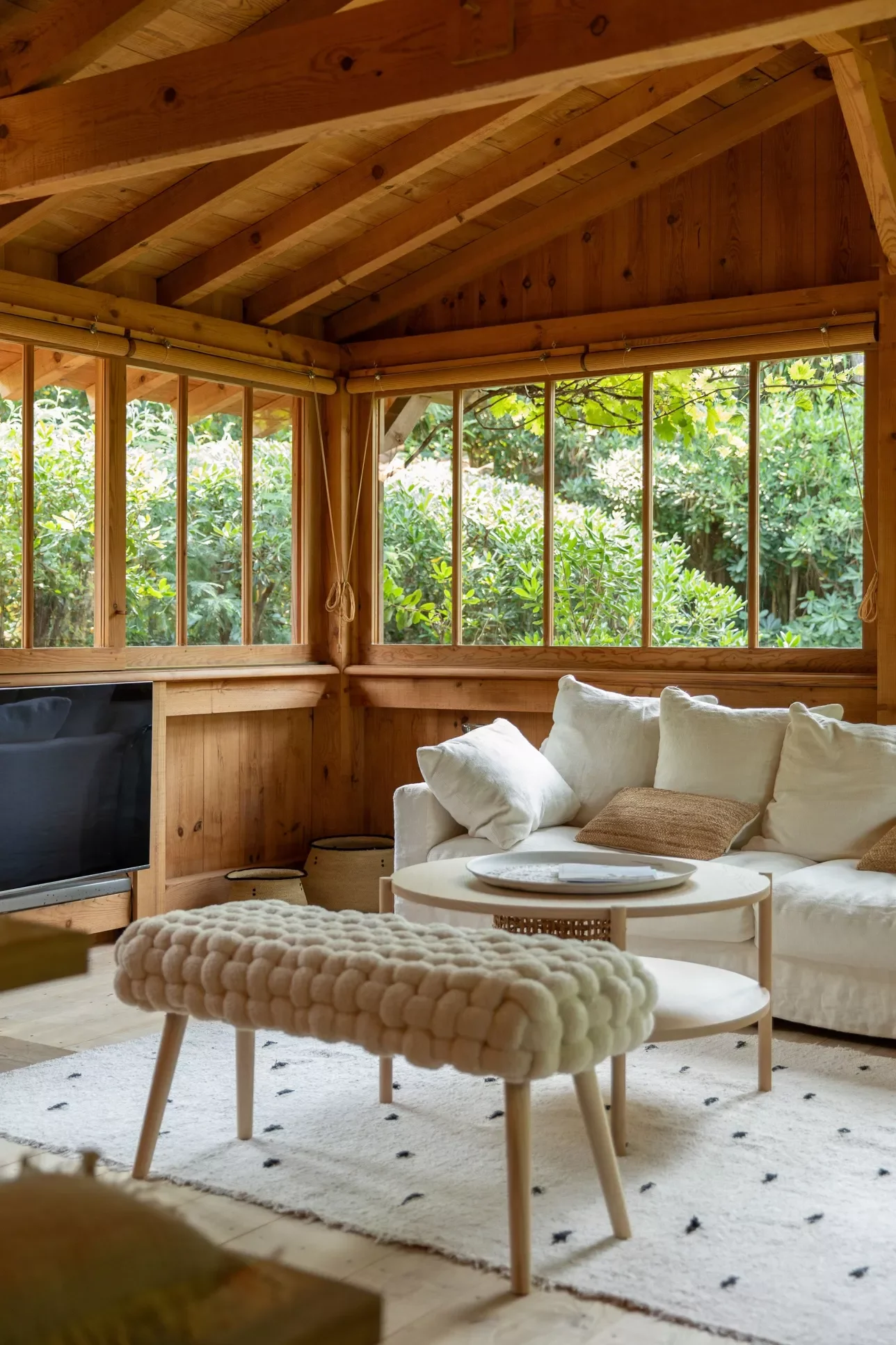 Salon cosy avec canapé blanc, fauteuil en tissu texturé, table basse ronde, tapis blanc avec petits motifs noirs, fenêtres avec vue sur la verdure extérieure, intérieur en bois.