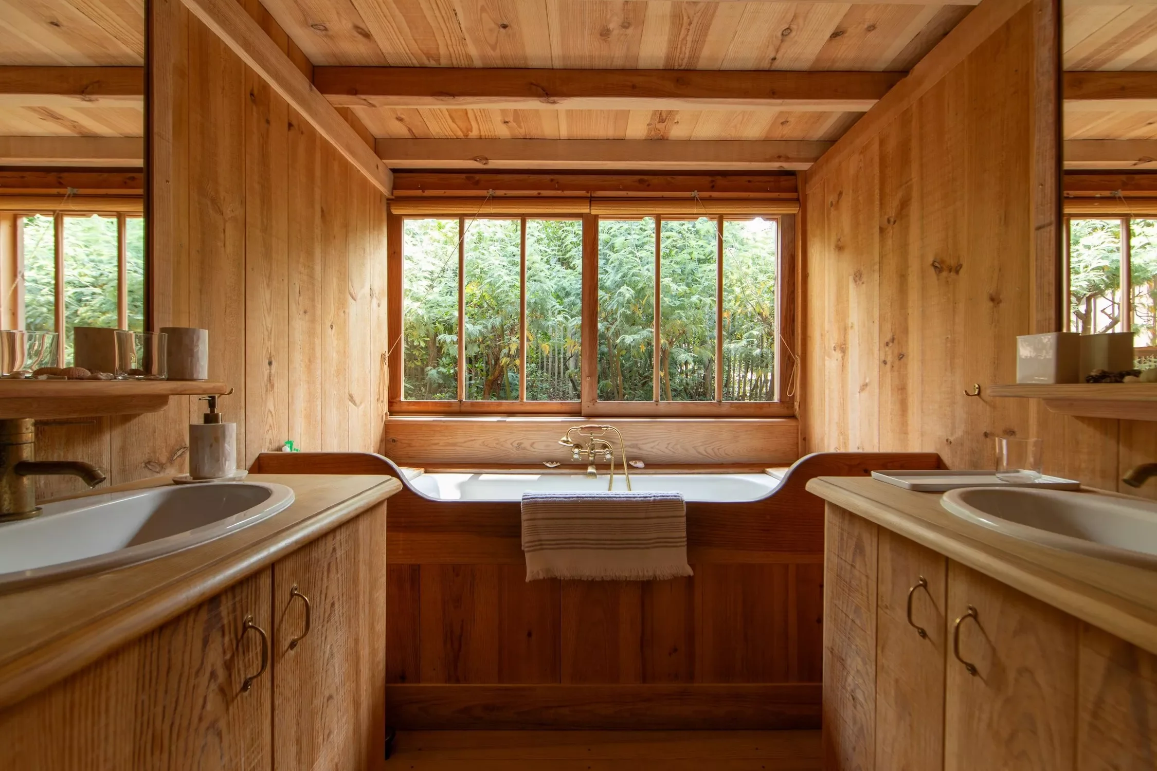 Salle de bain en bois avec double lavabo, grande fenêtre donnant sur un jardin vert, baignoire au centre.