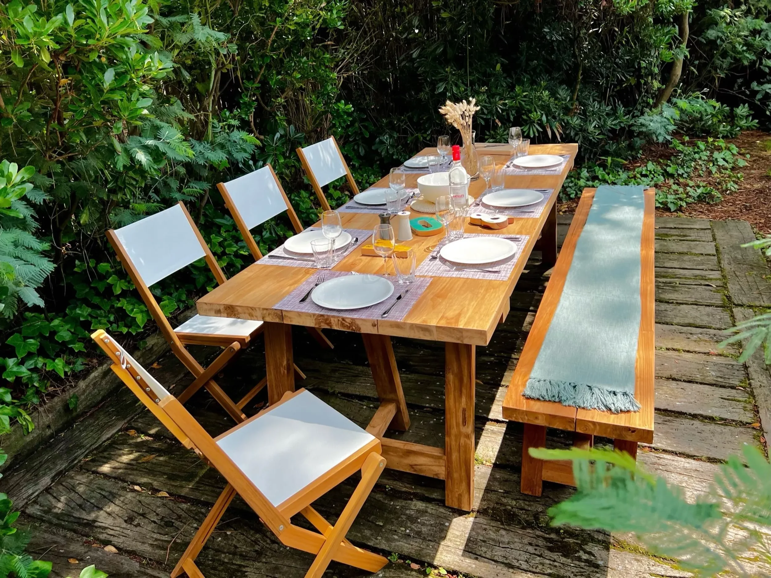 Une table en bois pour un repas en plein air est dressée avec des assiettes, des verres, et des serviettes, entourée d'herbes et vue sous un soleil doux.