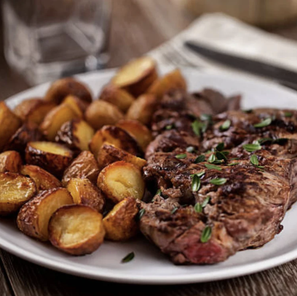 A black plate with cooked broccoli and a tomato-based vegetable dish on a wooden table. Nearby is a plastic container labeled 'Organic Chicken Cacciatore with Broccoli,' a glass with a green leafy plant, and a paper napkin with a logo that says 'MEMBERS Health Co'.