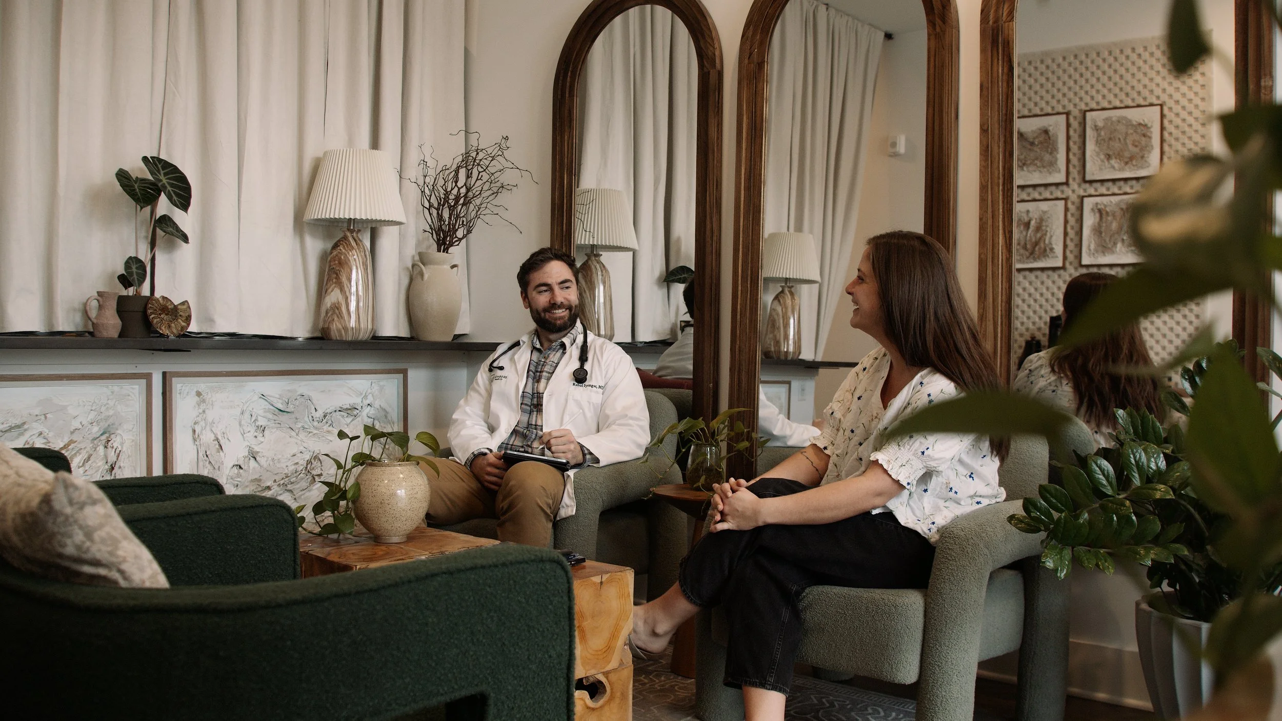 A male doctor in a white coat talking to a woman in black clothing, sitting in a cozy room with a mirror, plants, and decorative vases.