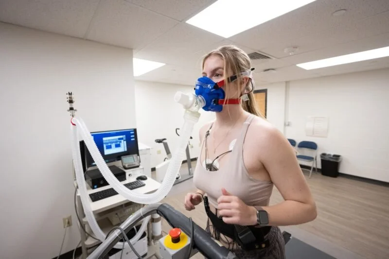 Woman on a treadmill wearing a mask connected to a machine in a medical or research setting.