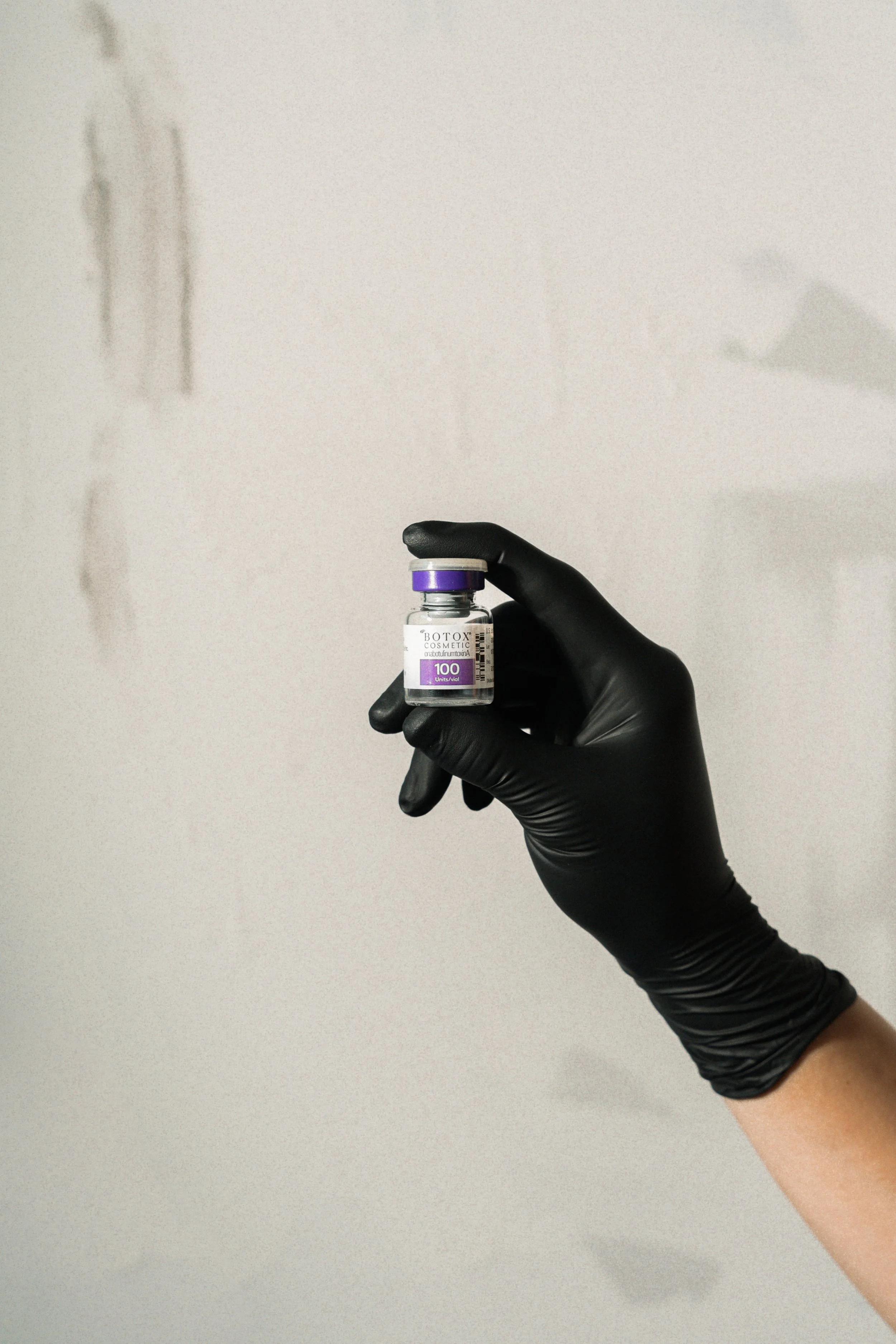A hand wearing a black glove holding a small vial labeled 'Botox Cosmetic' with purple accents.