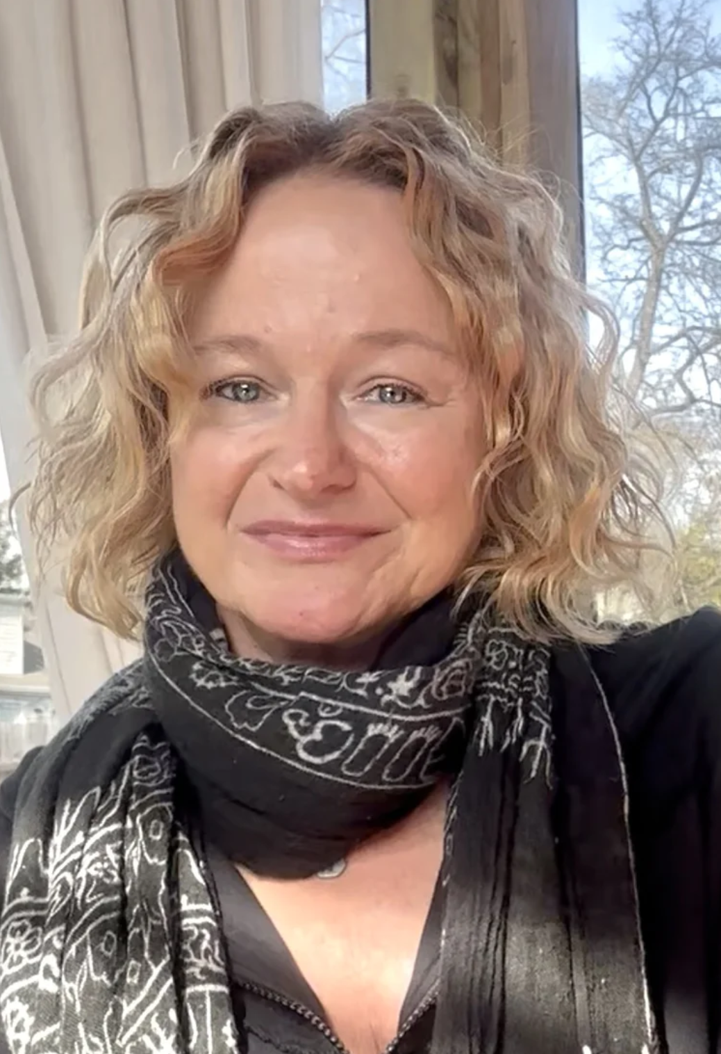 Close-up of a smiling woman with curly blonde hair, wearing a black scarf with white patterns and a black top, seated indoors with a window showing leafless trees outside.