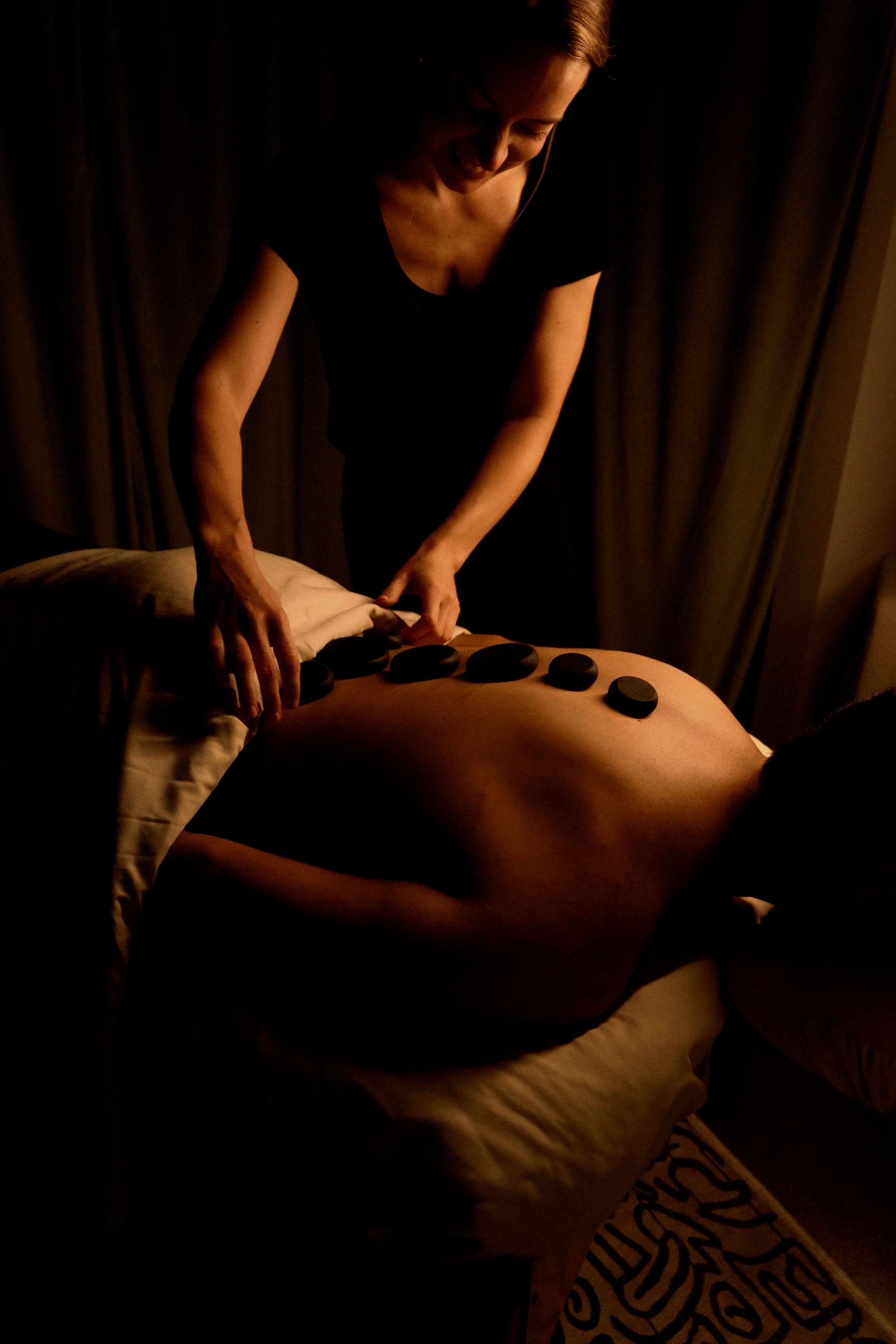 Close-up of a smiling woman with curly blonde hair, wearing a black scarf with white patterns and a black top, seated indoors with a window showing leafless trees outside.