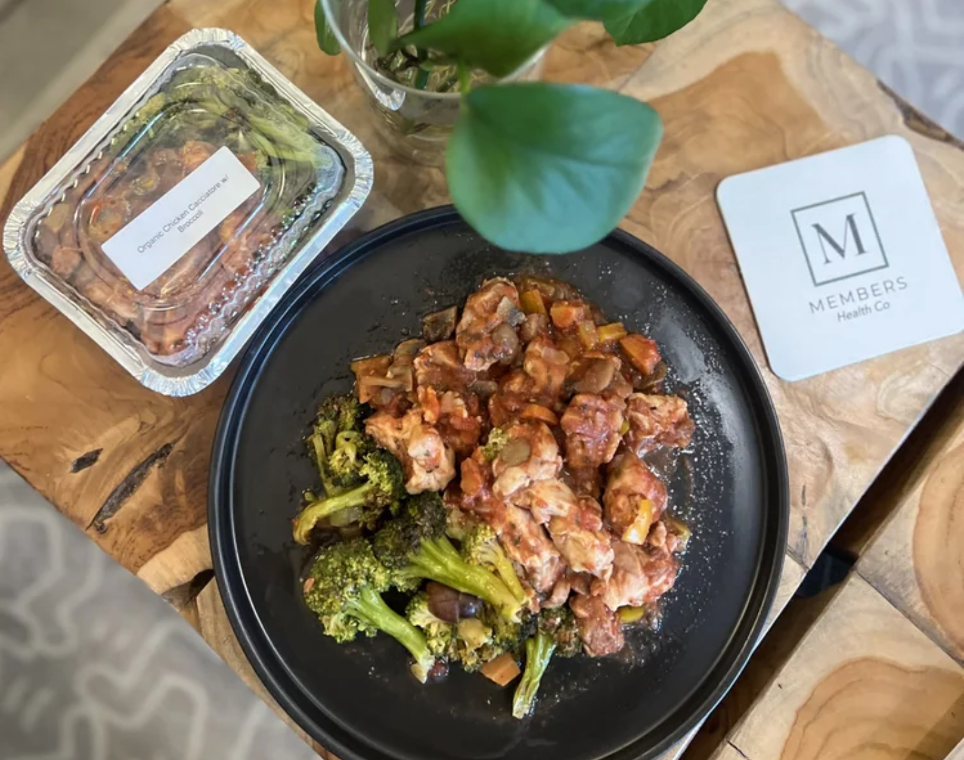 A black plate with cooked broccoli and a tomato-based vegetable dish on a wooden table. Nearby is a plastic container labeled 'Organic Chicken Cacciatore with Broccoli,' a glass with a green leafy plant, and a paper napkin with a logo that says 'MEMBERS Health Co'.