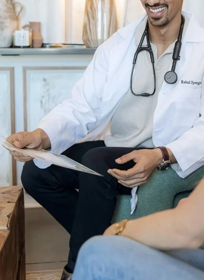 A male doctor with a stethoscope around his neck, wearing a white coat, sitting and holding a clipboard while speaking to a patient in a medical office or clinic.