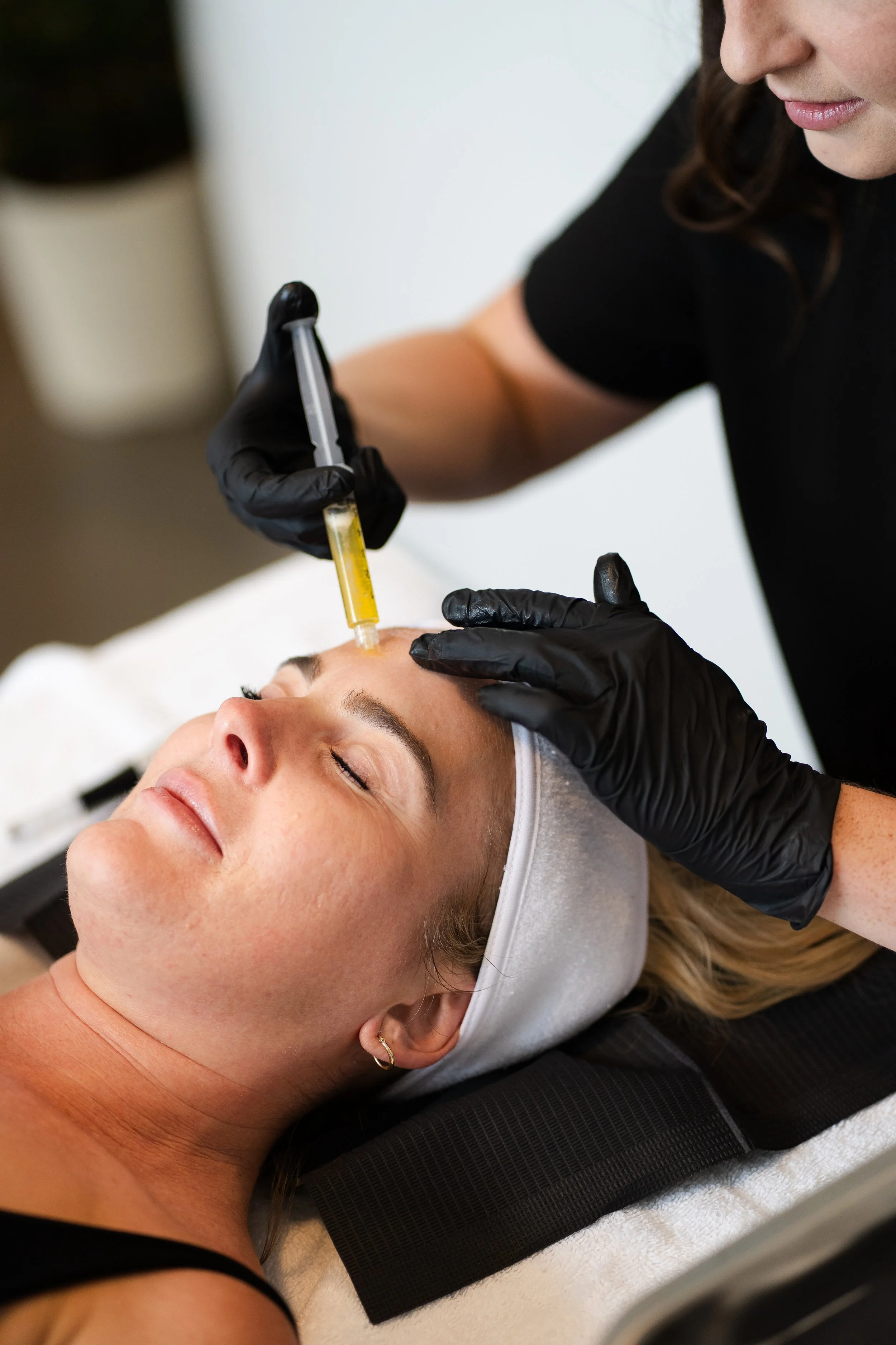 A woman receiving a facial treatment in a spa or clinic setting, lying on a treatment bed with a headband, while an aesthetician in black gloves injects a substance into her forehead with a syringe.