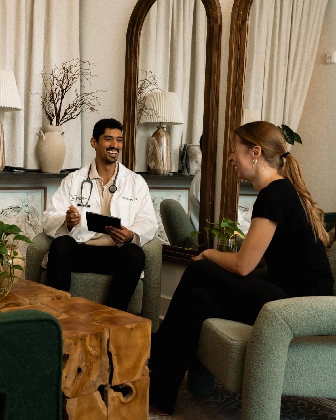 A doctor in a white coat and a woman with a black outfit having a conversation in a cozy room with a large mirror, plants, and lamps.