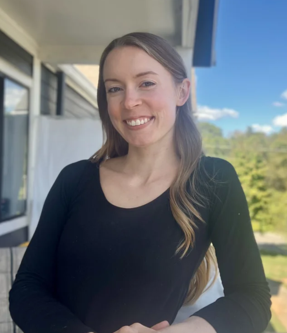 A woman smiling outdoors on a balcony with blue sky and green trees in the background.