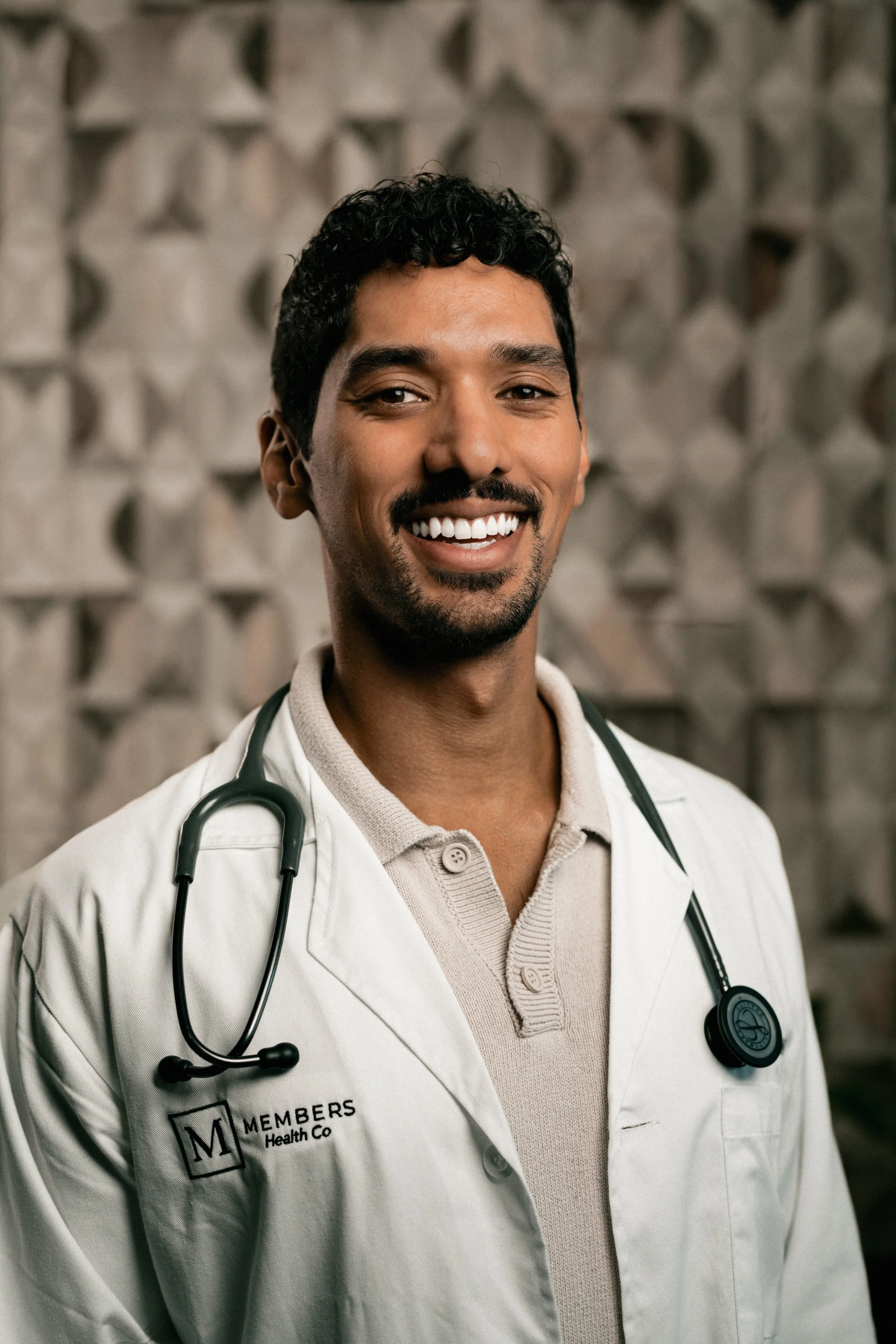 A smiling male doctor with a stethoscope around his neck, wearing a white lab coat with the logo 'Members Health Co.' in front of a patterned wall.