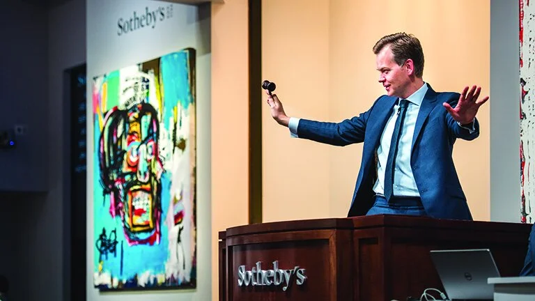 A man in a suit stands behind a Sotheby's auction podium, holding a microphone and raising his hand, with a vibrant colorful artwork displayed on a screen behind him.